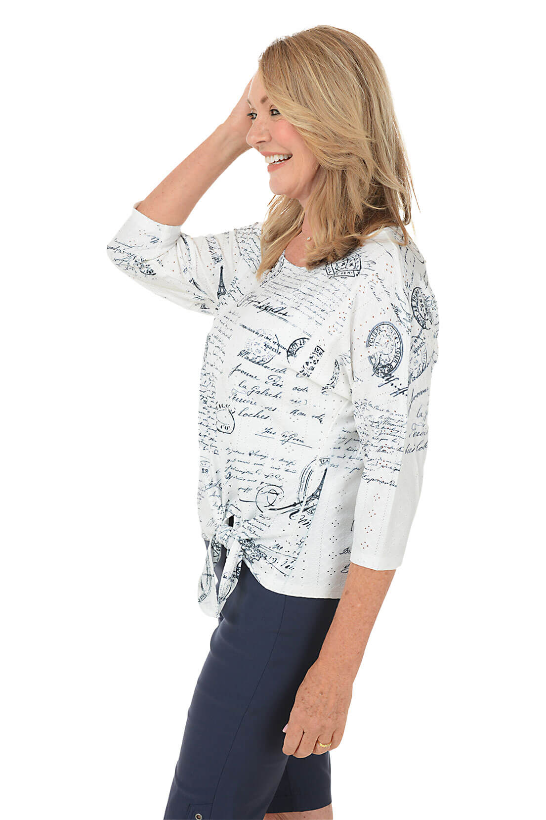 A side shot of a model wearing a petite white knit top with text pattern, 3/4 dolman sleeves, and a tie-front hemline. The top features a scoop neck and eyelet details.