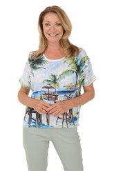 A front shot of a model wearing a white scoop neck knit top with short dolman sleeves and sequined cuffs, featuring a beach bar print.