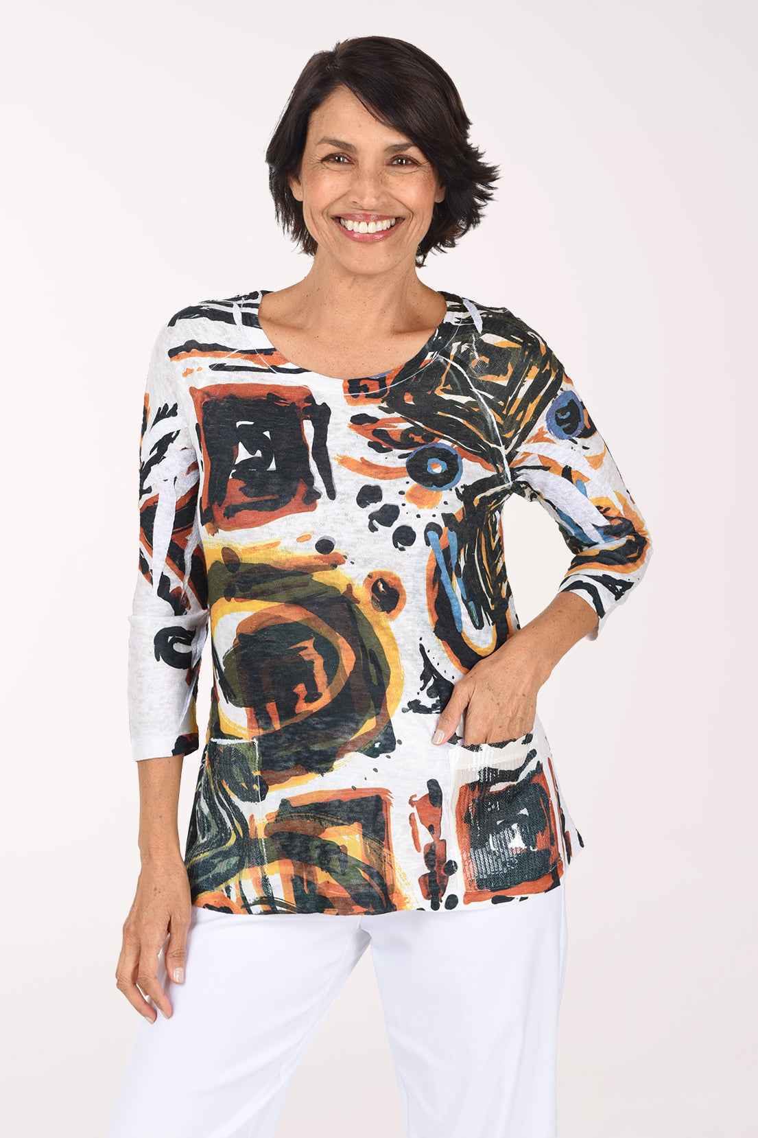 Smiling woman in a white semi-sheer scoop neck knit top with a bold abstract print of amber, rust, olive and black spirals and squares, worn with white pants