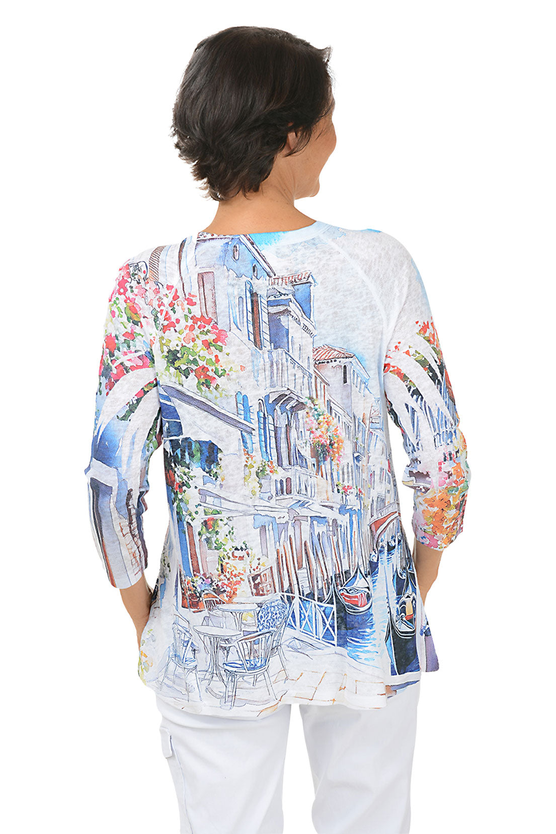 Woman wearing a colorful venice patterned 3/4 sleeve top with white pants on a white background. Back shot.