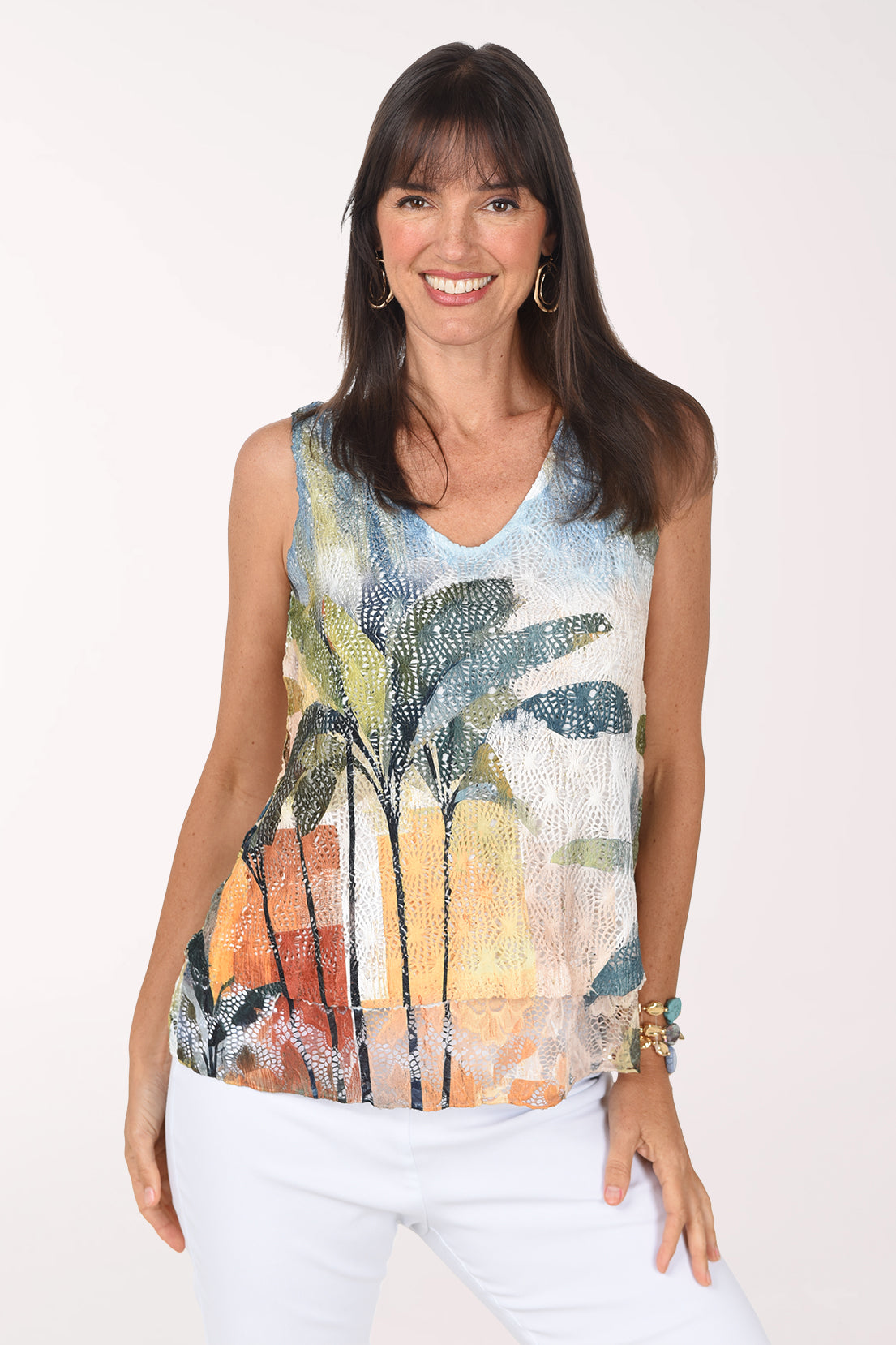 Front view of a woman in a sleeveless V-neck crochet top printed with painterly green and teal palm trees against an orange, rust and golden sunset, worn with white jeans.