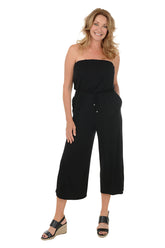 A front shot of a model standing, wearing a black strapless wide leg jumpsuit with an elastic top band and adjustable drawstring waist, with front pockets. She is also wearing a pair of black wedges.