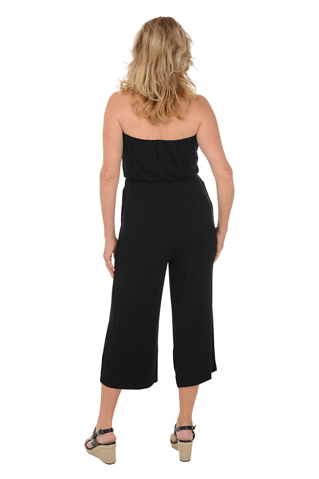 A back shot of a model standing, wearing a black strapless wide leg jumpsuit with an elastic top band and adjustable drawstring waist, with front pockets. She is also wearing a pair of black wedges.