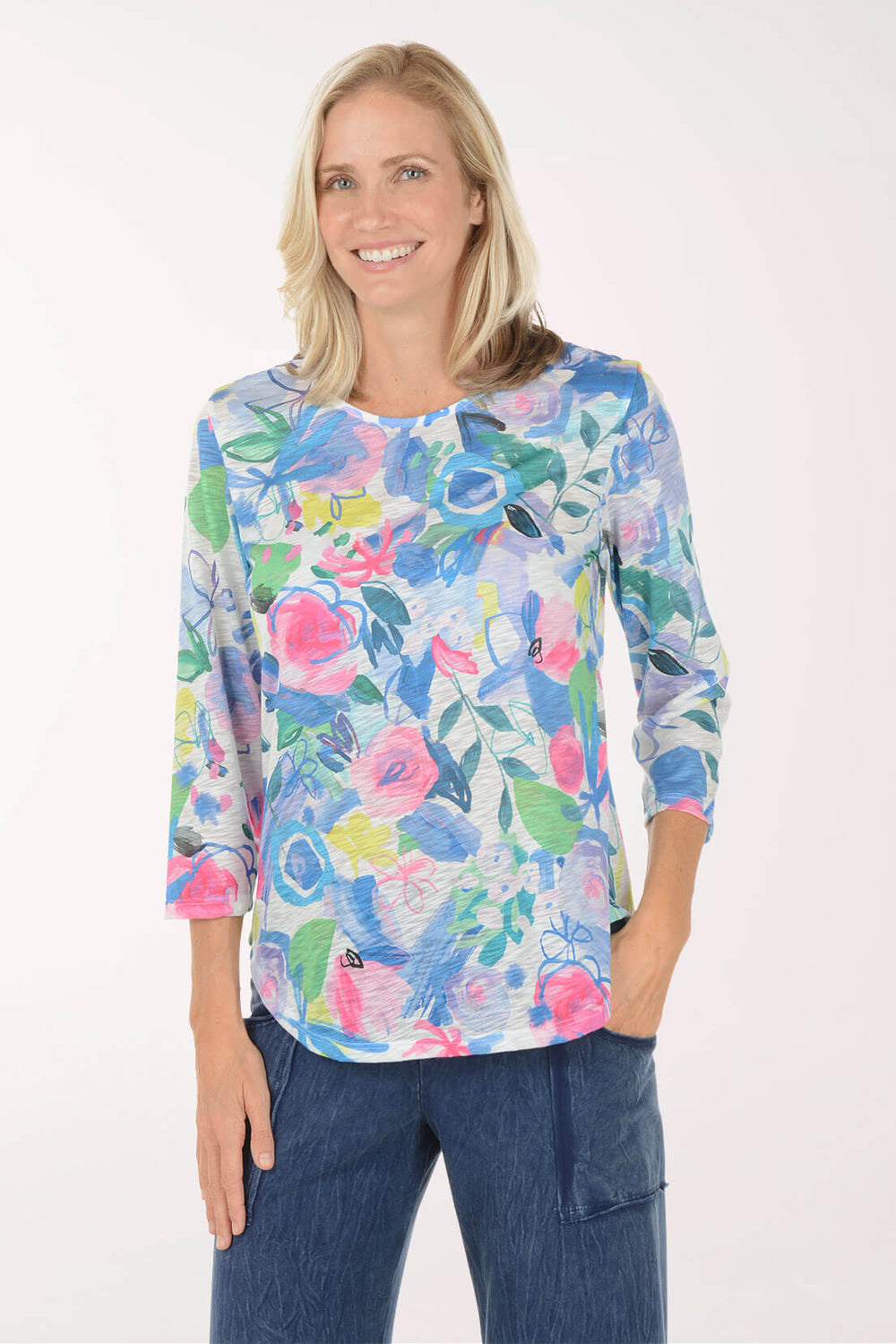 Woman smiling with one hand in her pocket, wearing a white slub knit top with a painterly floral print in cobalt and periwinkle blue, hot pink, green, yellow, and lavender, with a scoop neckline, 3/4 sleeves, and a curved hem, worn with dark wash wide leg jeans.