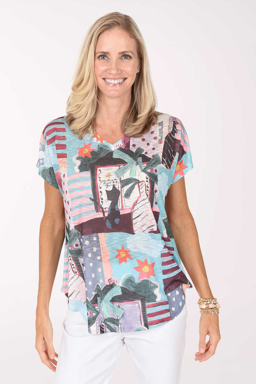 Model wearing the Jess and Jane Sunny Day Slub Knit Short Sleeve Top, a pastel patchwork of palms and beach scenes, over white pants, front view