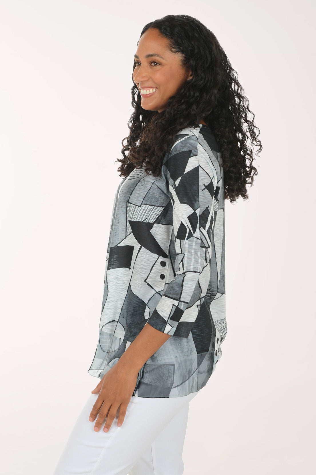 A woman with long curly dark hair wearing a loose-fitting, three-quarter sleeve top with an abstract geometric print in black, gray, and white, paired with white pants, against a plain light background.