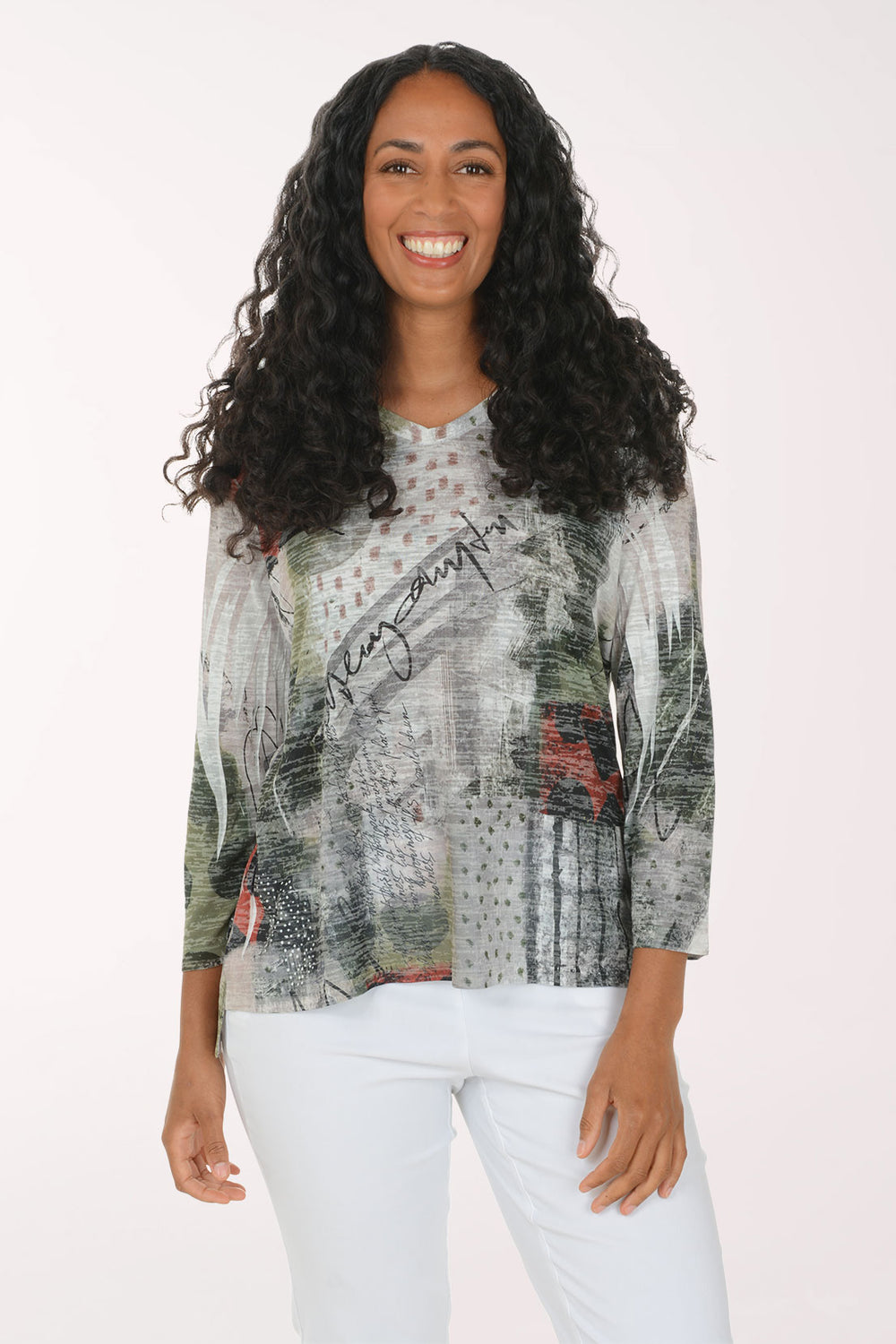 A woman with long, curly black hair is smiling and wearing a three-quarter sleeve top with an abstract, painterly print in gray, green, black, and red, paired with white pants, against a plain white studio background.