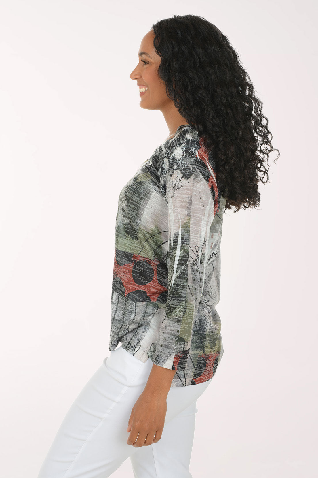 A woman with long, curly black hair is smiling and wearing a three-quarter sleeve top with an abstract, painterly print in gray, green, black, and red, paired with white pants, against a plain white studio background.