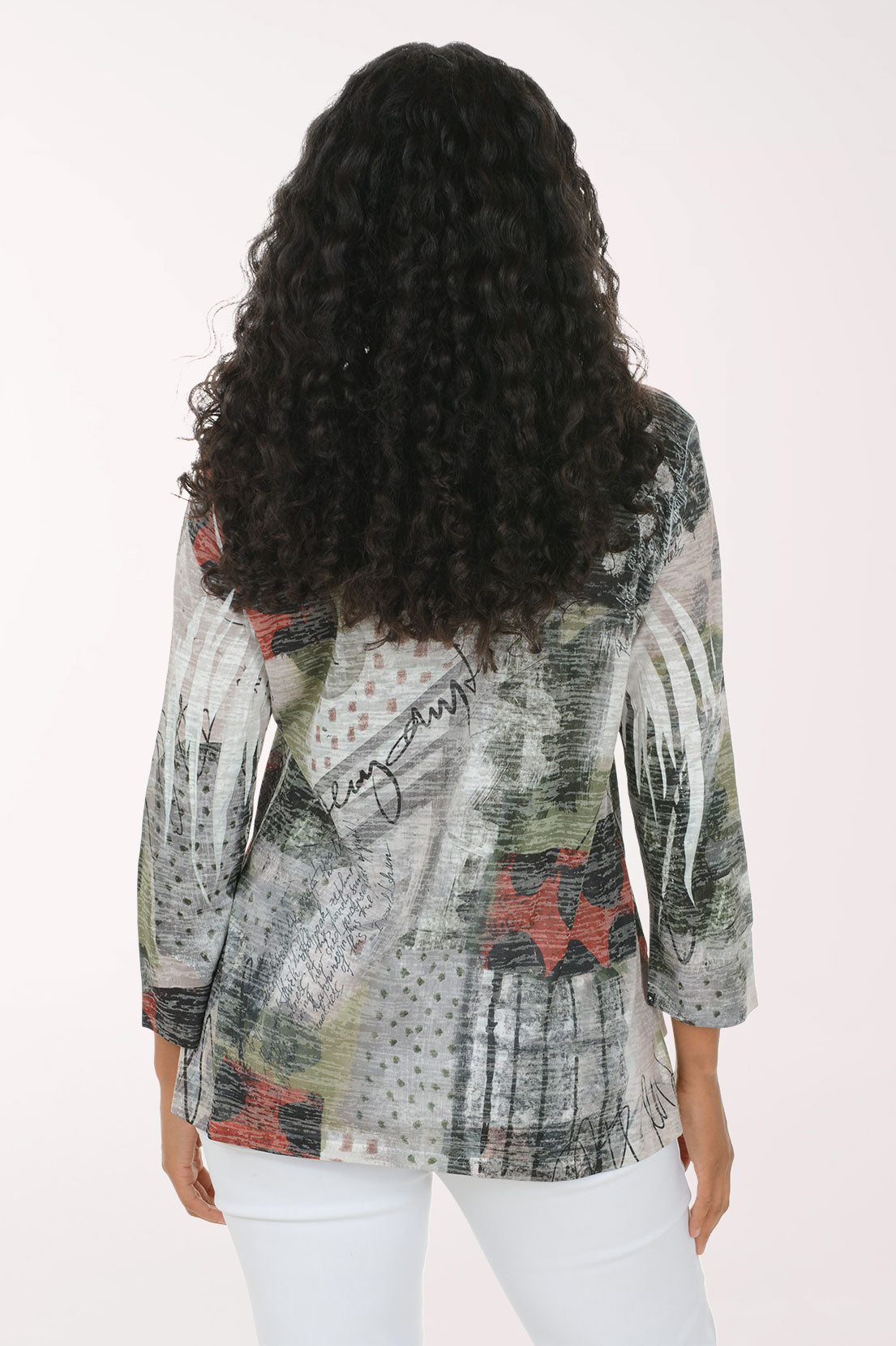 A woman with long, curly black hair is smiling and wearing a three-quarter sleeve top with an abstract, painterly print in gray, green, black, and red, paired with white pants, against a plain white studio background.