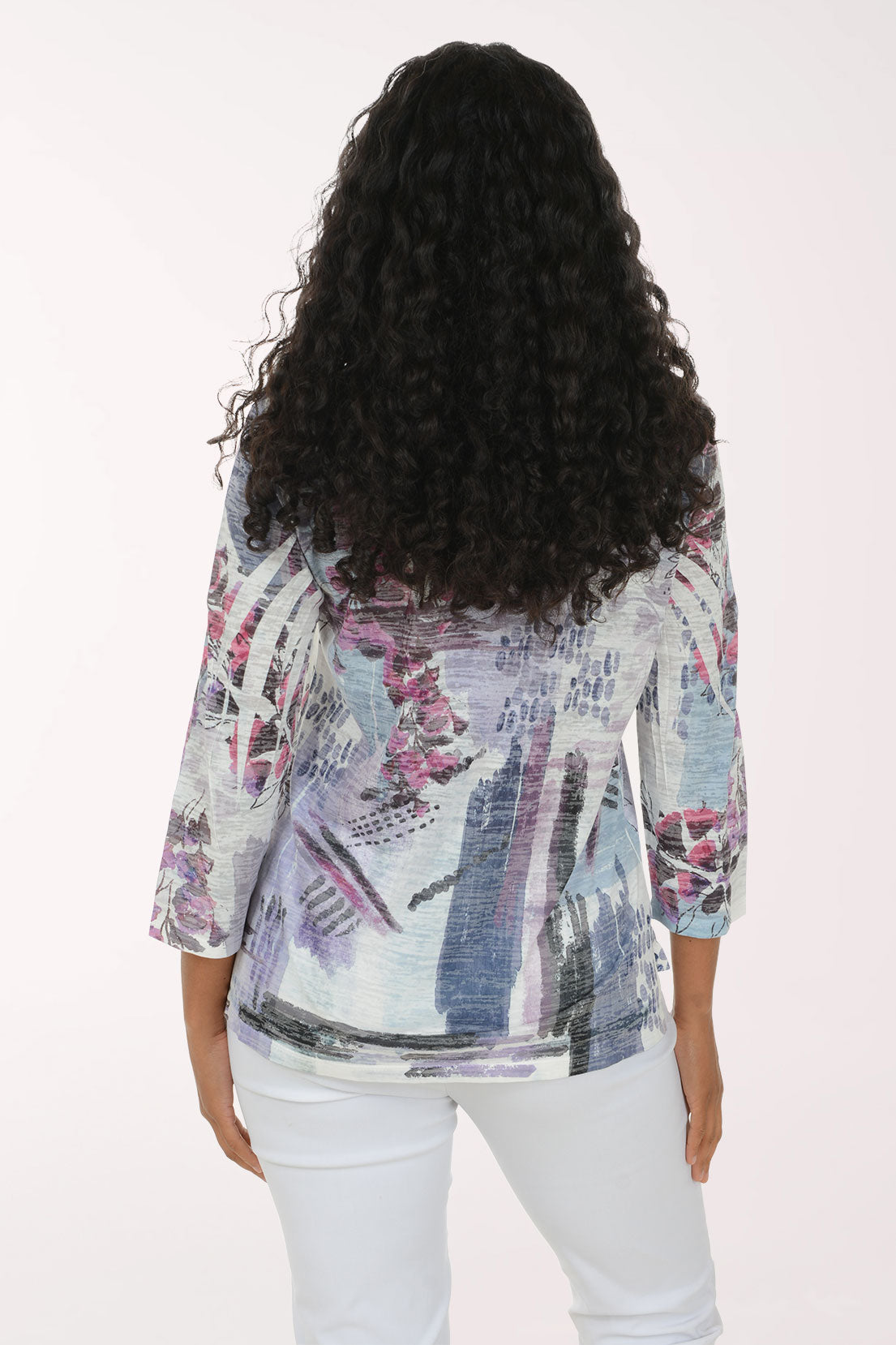 A woman with long, curly black hair wearing a three-quarter sleeve blouse with a blue, purple, and pink abstract botanical print, paired with white pants, standing against a plain white studio background.