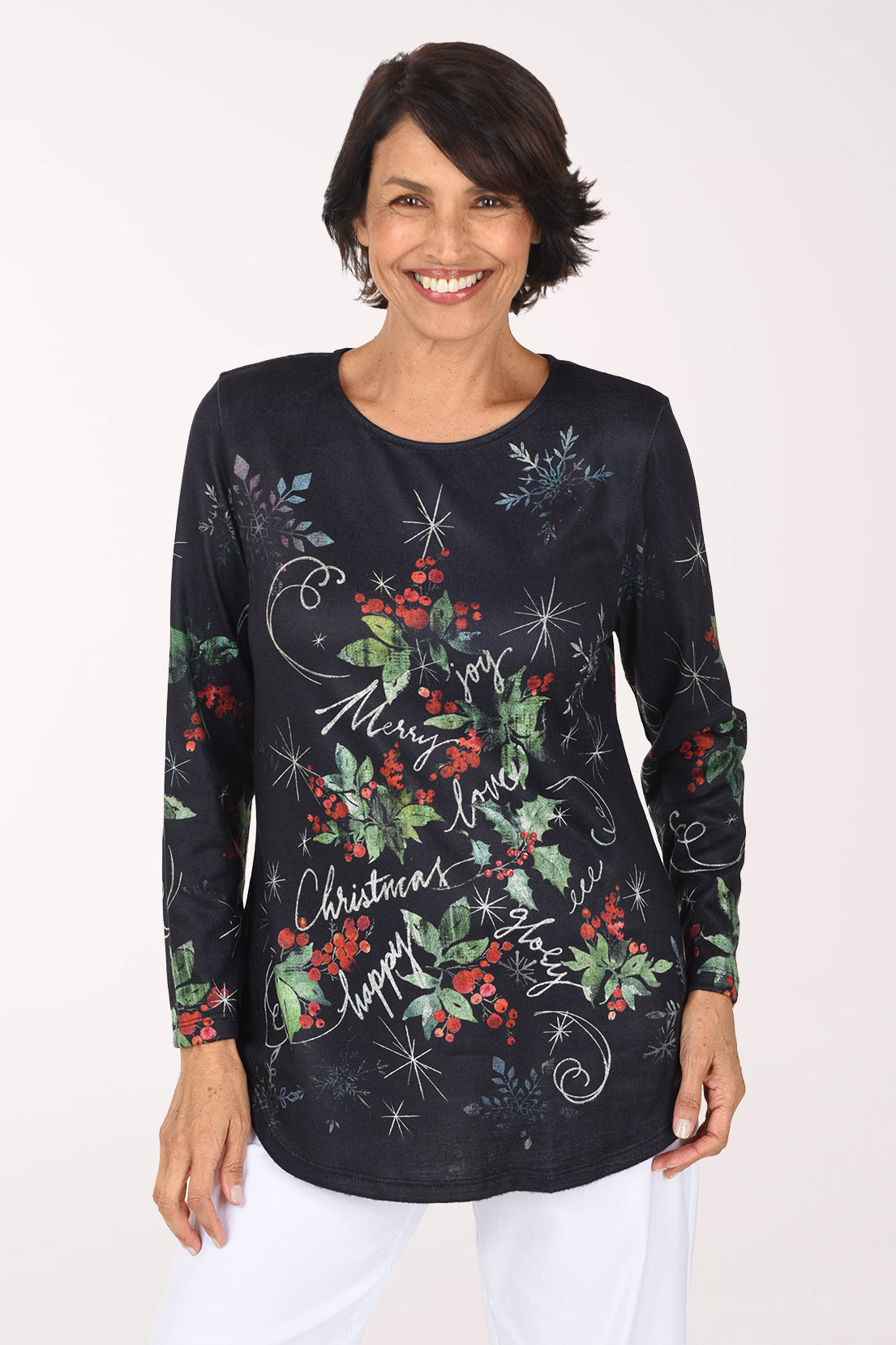 Smiling woman in a black long sleeve scoop neck top with an all-over print of red holly berries, green leaves, white starbursts and holiday script reading Merry, Joy and Christmas, worn with white pants