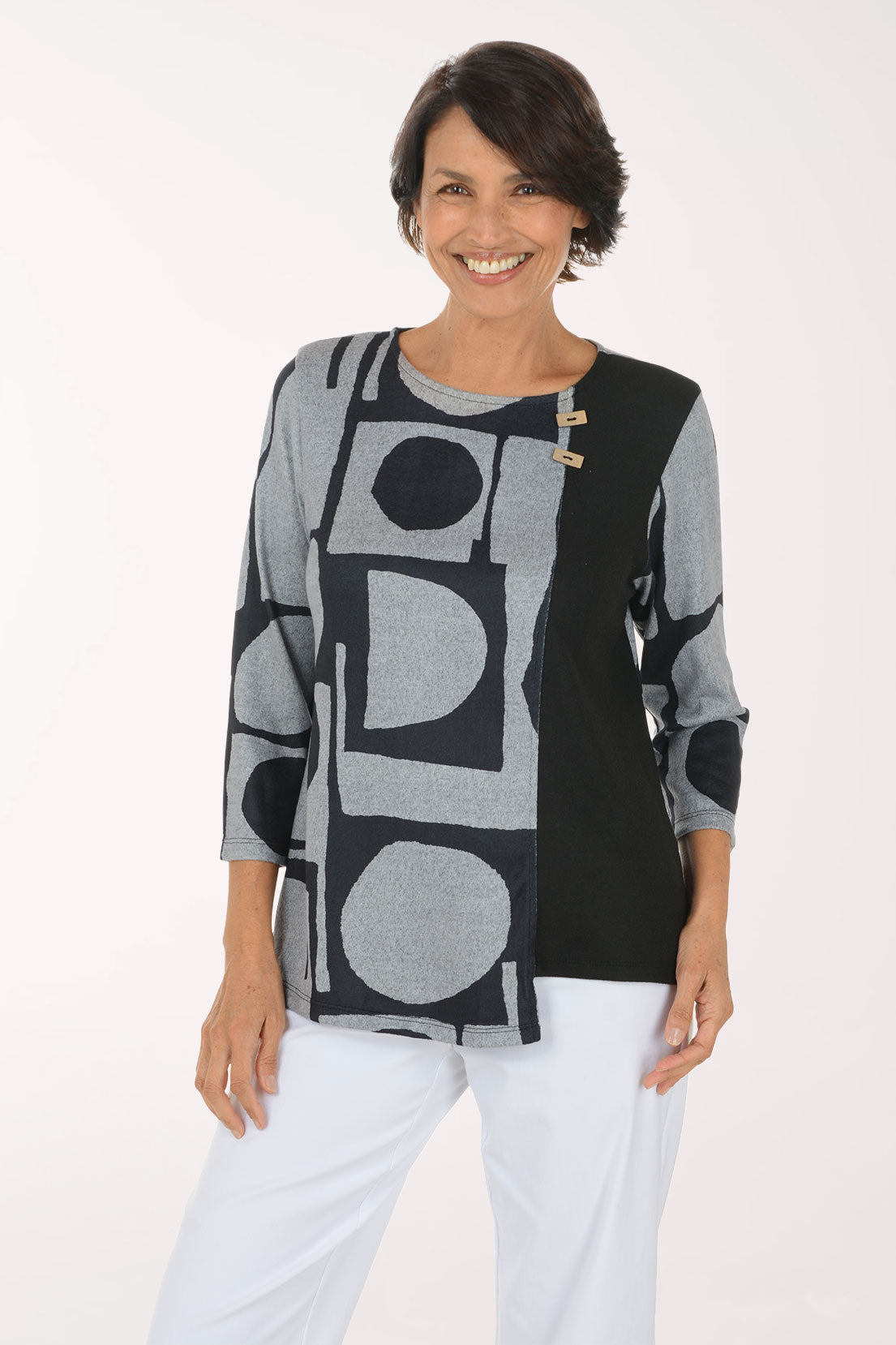 A smiling woman wearing a long-sleeve knit top with a black and gray geometric pattern, paired with white pants, photographed against a plain light background.