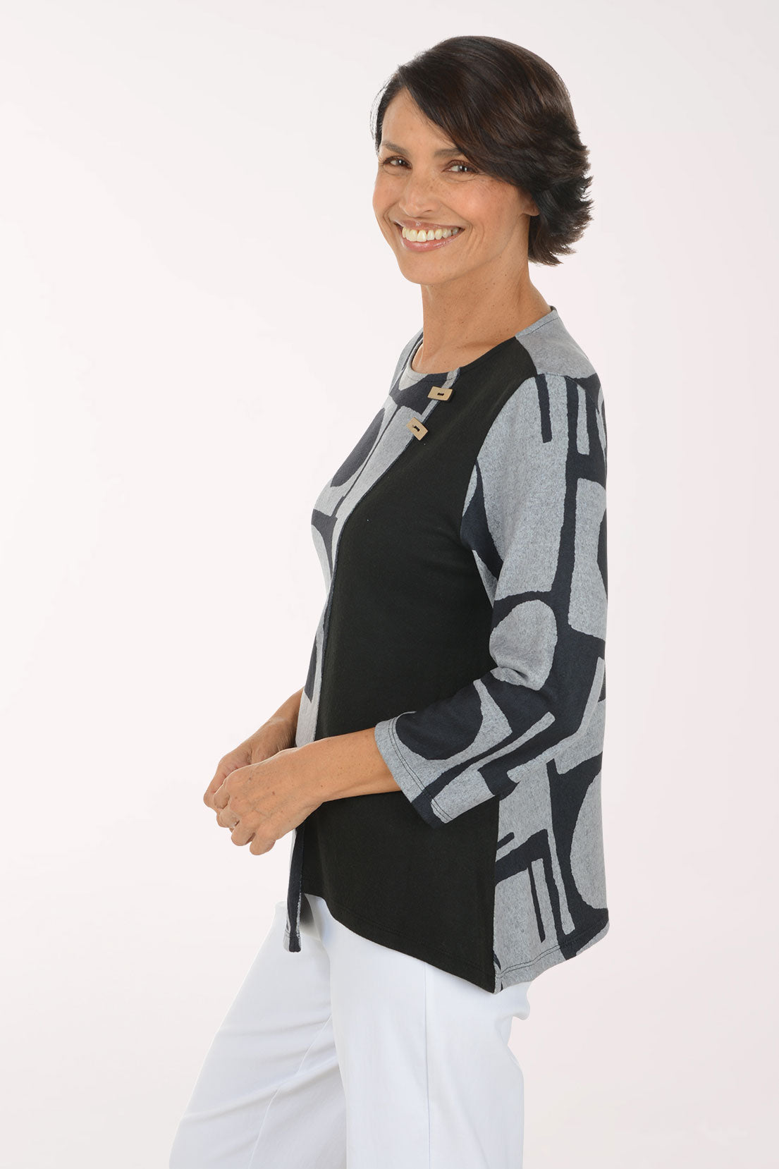 A smiling woman wearing a long-sleeve knit top with a black and gray geometric pattern, paired with white pants, photographed against a plain light background.