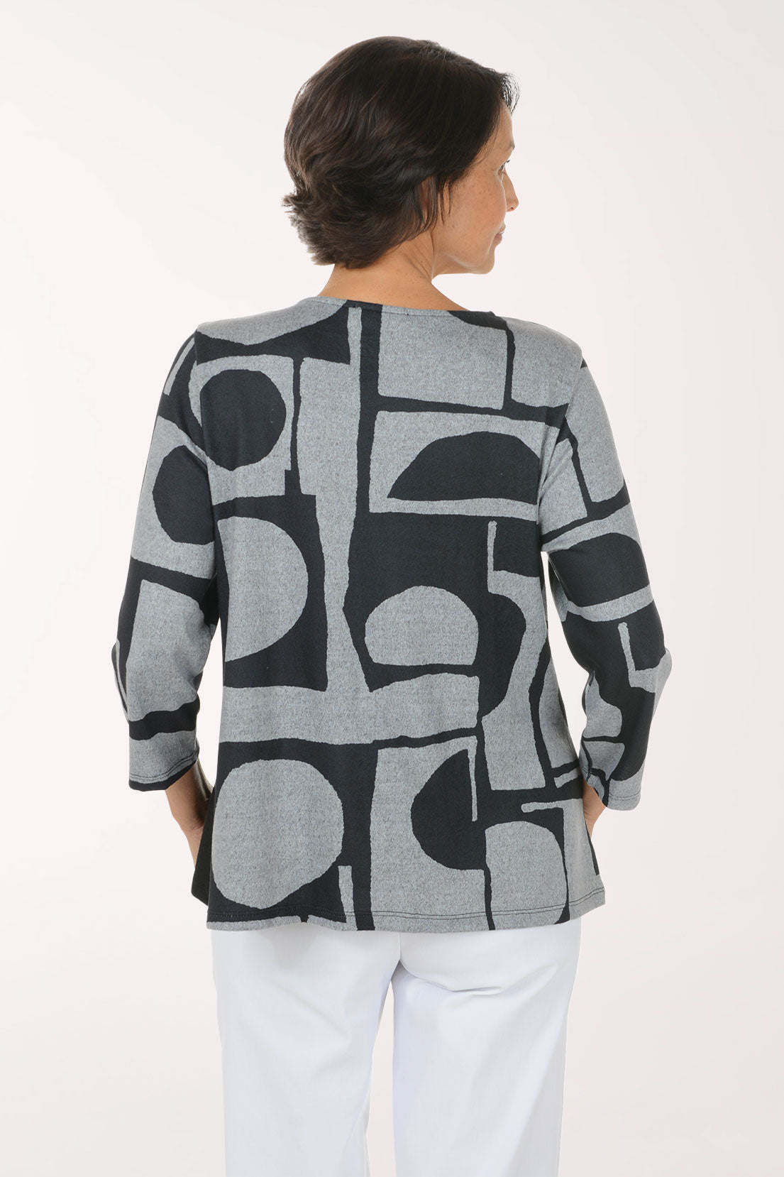 A smiling woman wearing a long-sleeve knit top with a black and gray geometric pattern, paired with white pants, photographed against a plain light background.