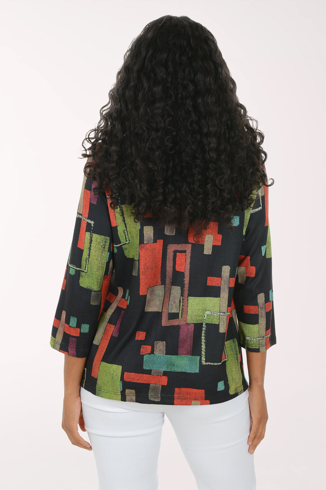 A smiling woman with long curly black hair wearing a black three-quarter sleeve top with a colorful geometric block pattern in red, green, and beige, paired with white pants, against a plain white studio background.
