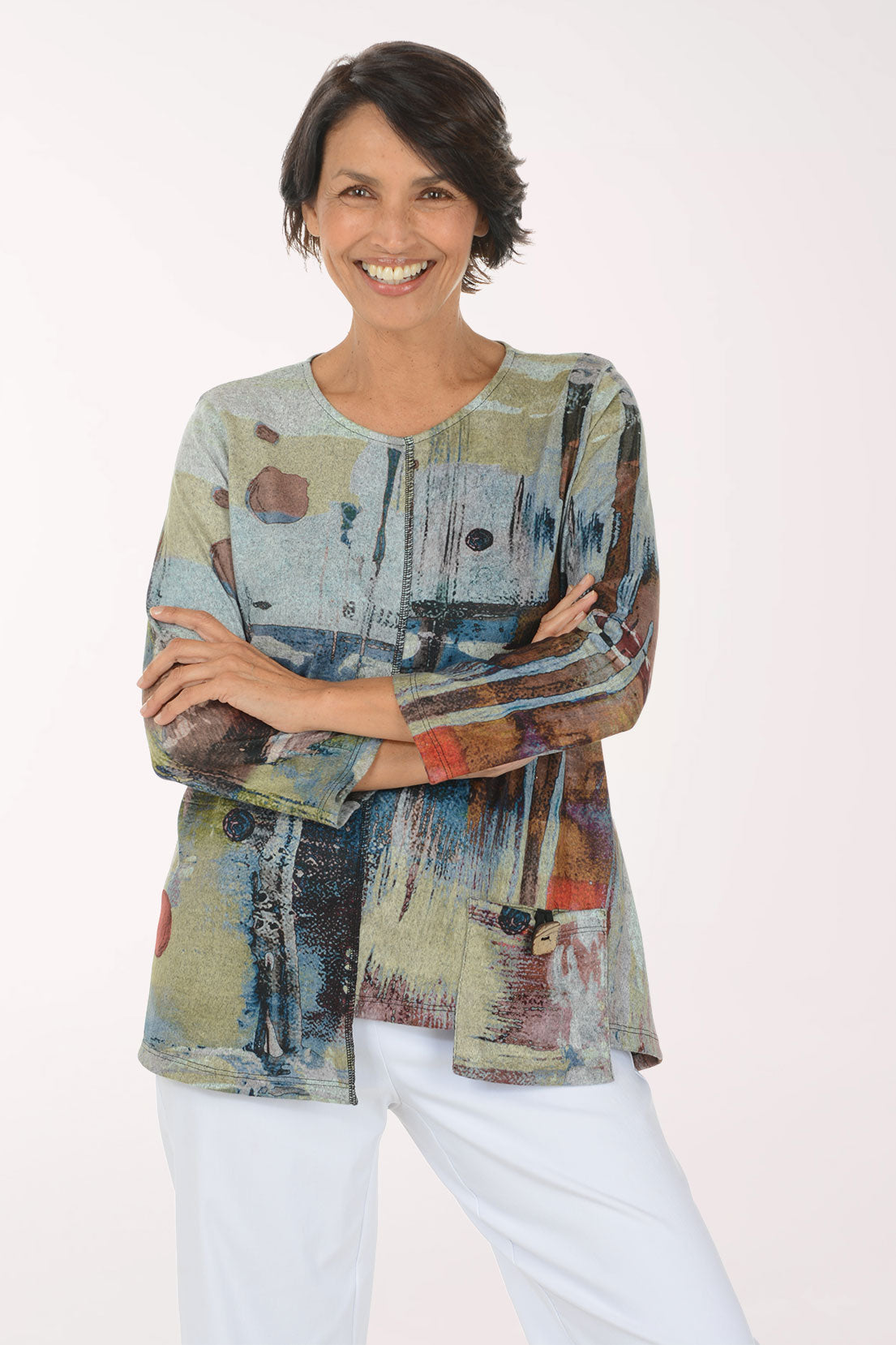 A woman modeling a long-sleeve abstract print top with blue, beige, brown, and red tones, paired with white pants, against a plain light background.