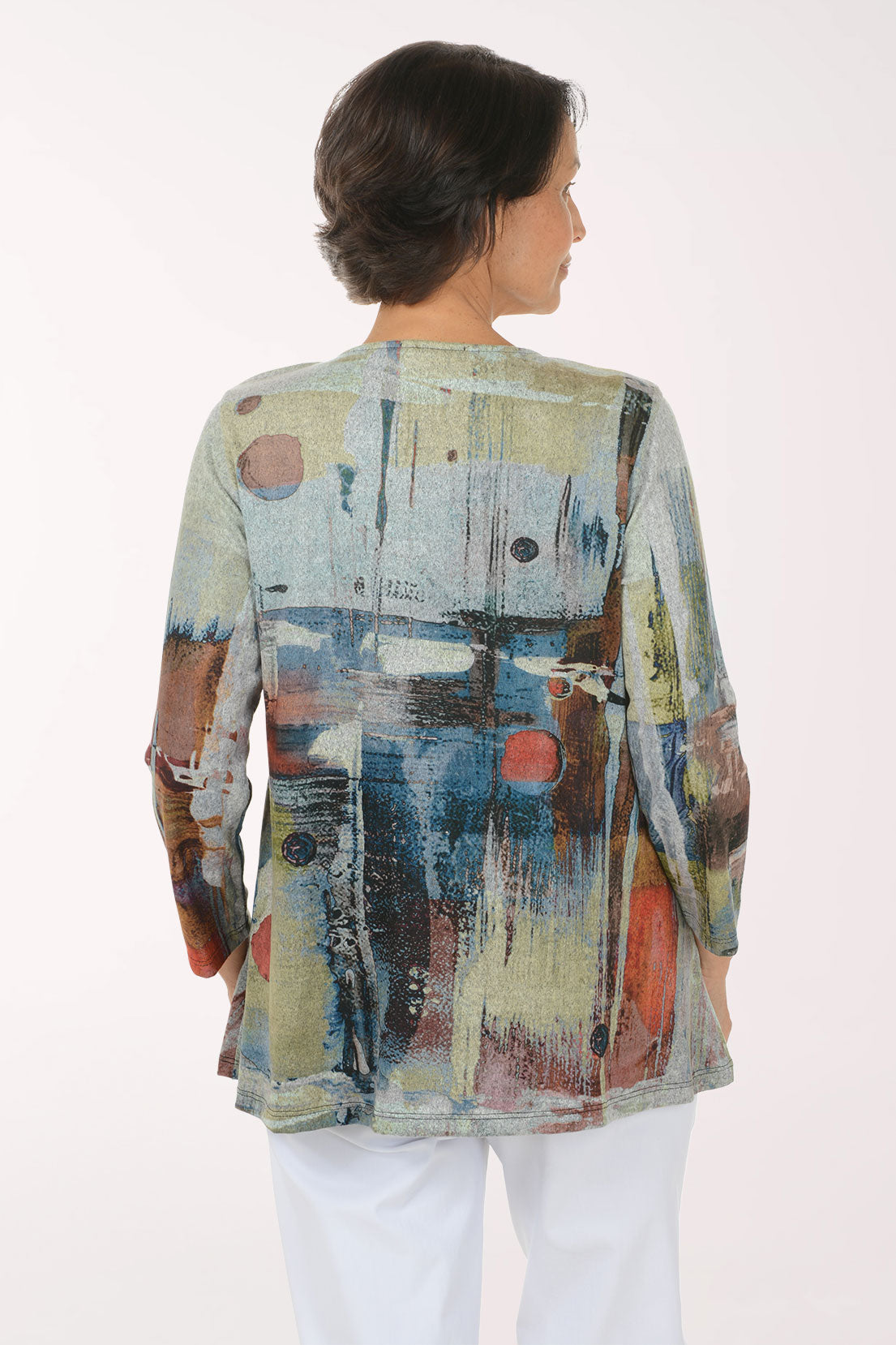 A woman modeling a long-sleeve abstract print top with blue, beige, brown, and red tones, paired with white pants, against a plain light background.