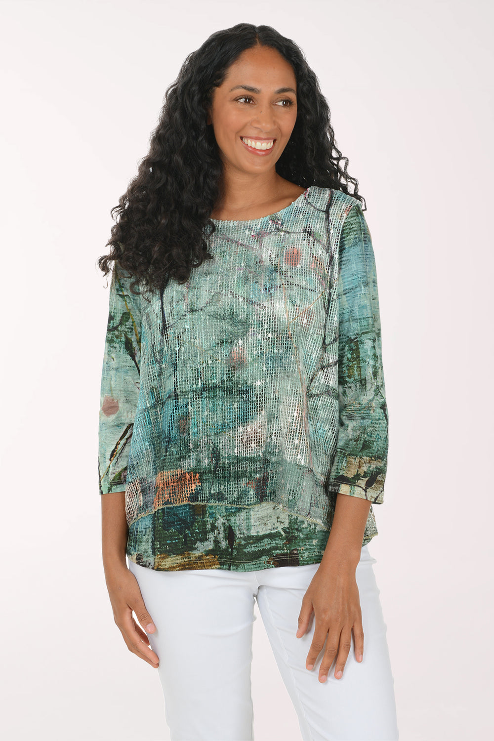 A woman with long, curly black hair wearing a green and blue abstract-patterned blouse and white pants, standing against a plain white studio background.