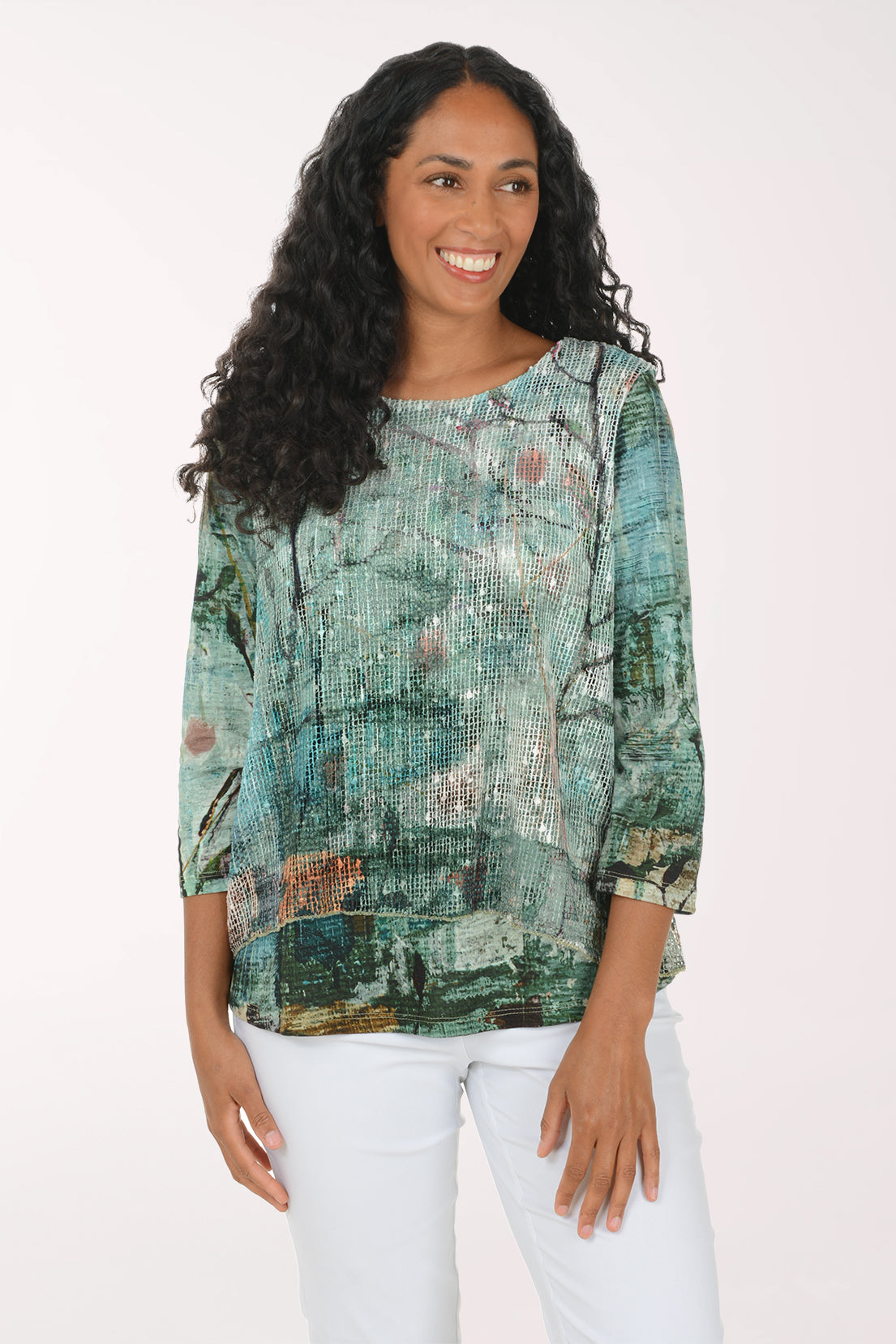 A woman with long, curly black hair wearing a green and blue abstract-patterned blouse and white pants, standing against a plain white studio background.