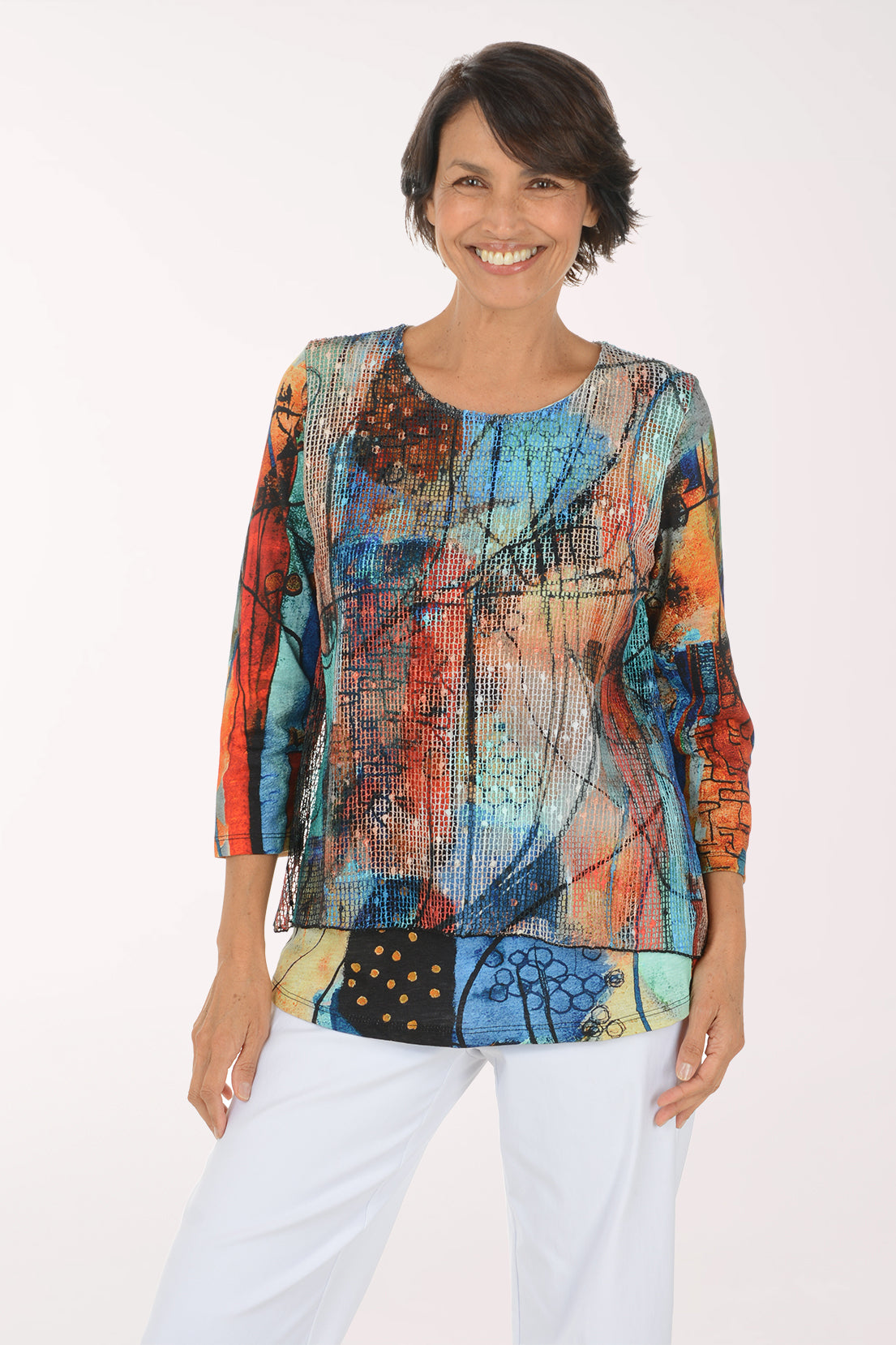 A smiling woman wearing a colorful abstract print top with three-quarter sleeves and white pants, standing against a plain white studio background.