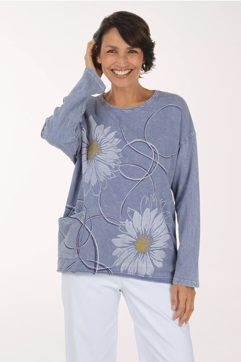 A smiling woman wearing a light blue long-sleeve top with large white daisy floral prints and white pants, standing against a plain white studio background.