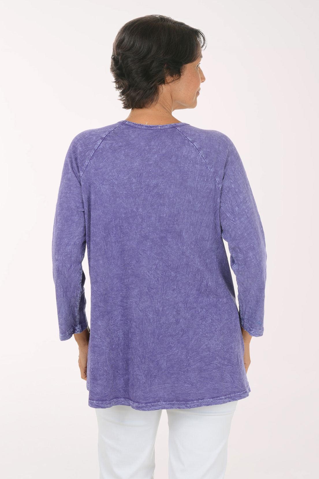 A smiling woman wearing a lavender-purple long-sleeve tunic top with a colorful abstract floral and botanical print, paired with white pants, against a plain white studio background.