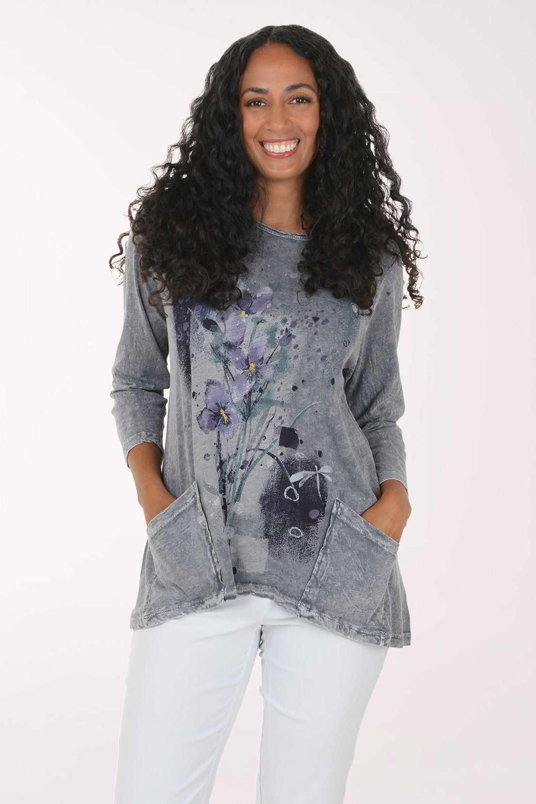 A woman with long, curly black hair wearing a gray mineral-wash long-sleeve tunic top with a purple and black floral print, paired with white pants, standing against a plain white background.