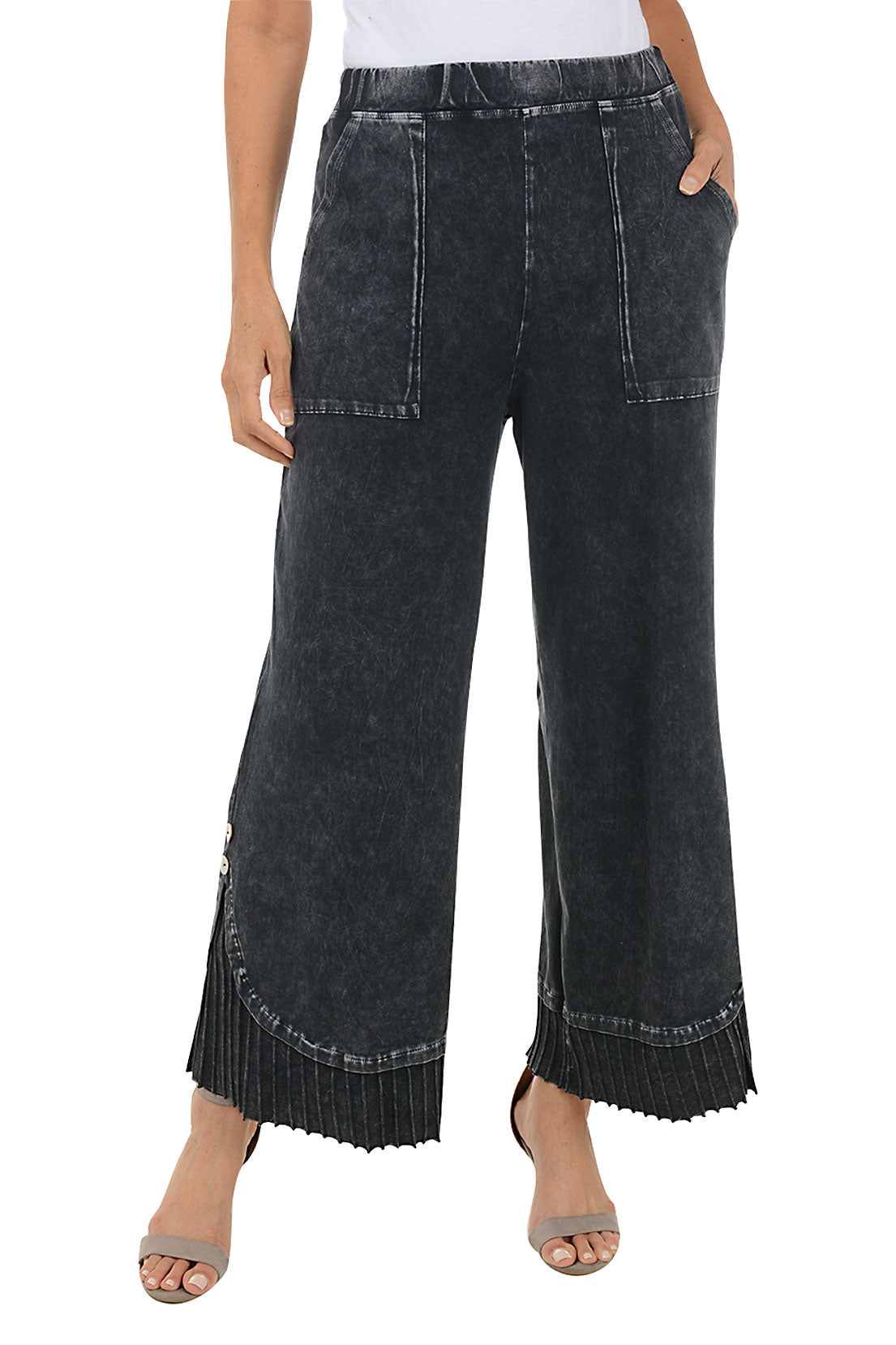 A front shot of a model wearing a pair of black denim wide-leg, pleated hem pants with a mineral wash design. The pants have a high waist with a 1 1/2 inch elasticized waistband, dual front patch pockets, and a double button embellishment at the pleated hemline.