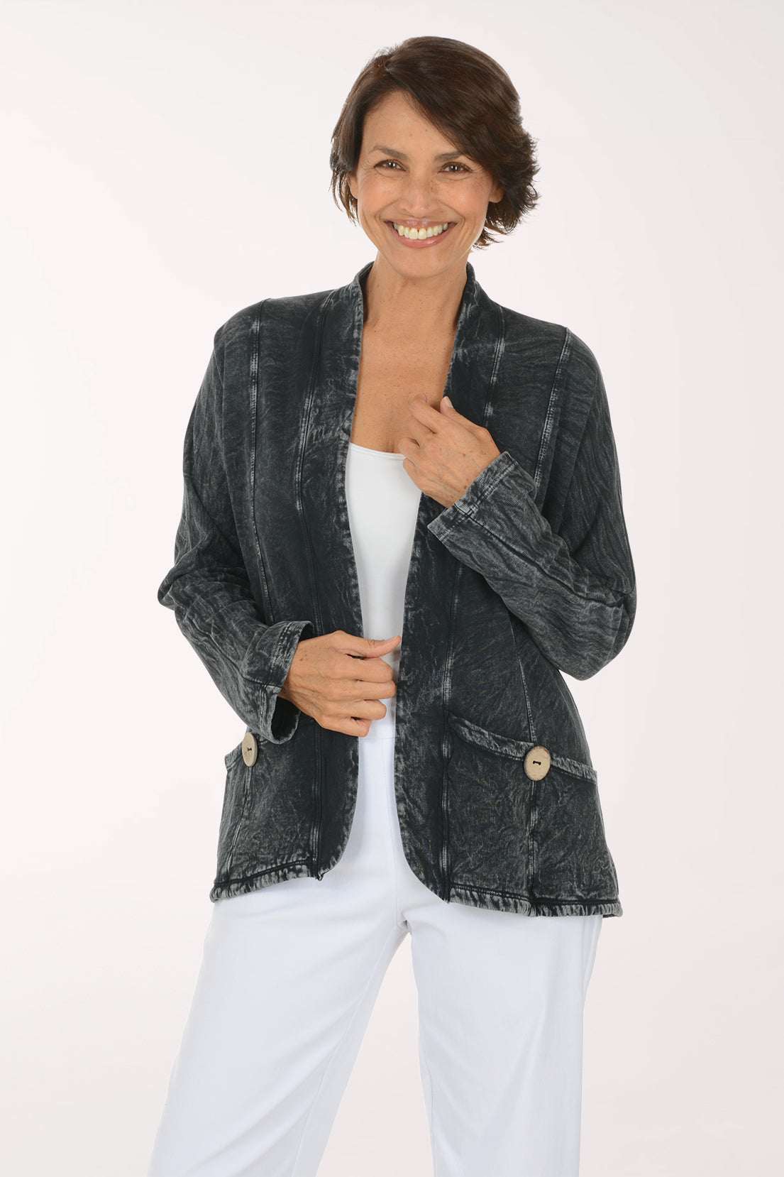 A smiling woman wearing a dark gray mineral-wash denim jacket over a white top and white pants, photographed against a clean white studio background.