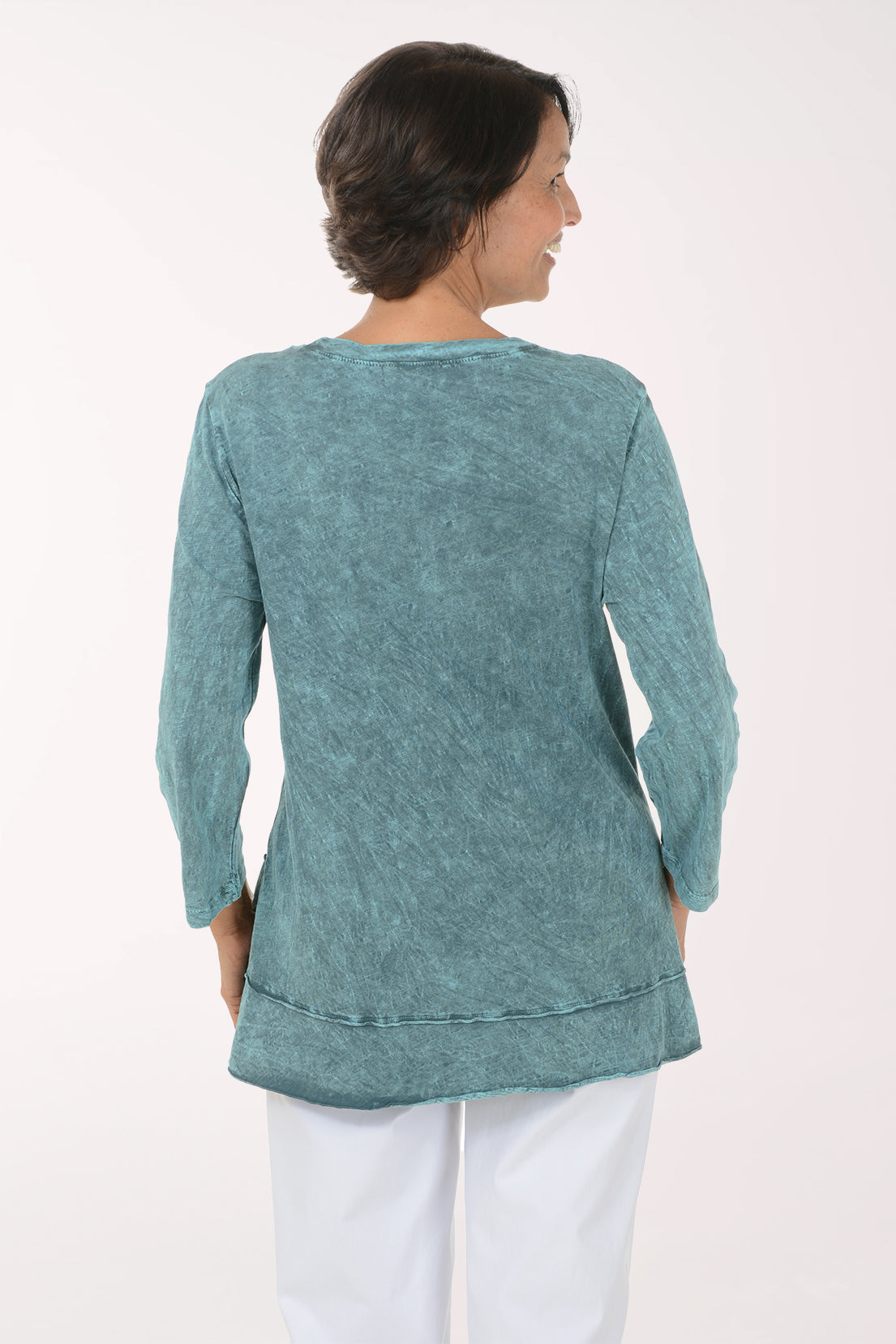 A smiling woman wearing a long-sleeve, tiered tunic top in a washed teal blue color with abstract black and red botanical line drawings, paired with white pants, against a plain white studio background.