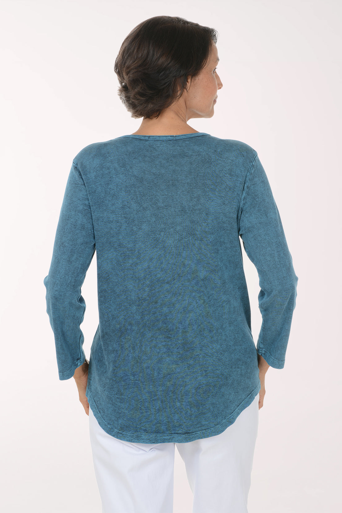 A smiling woman wearing a long-sleeve blue tunic top with a yellow and green floral print, paired with white pants, against a plain white background.