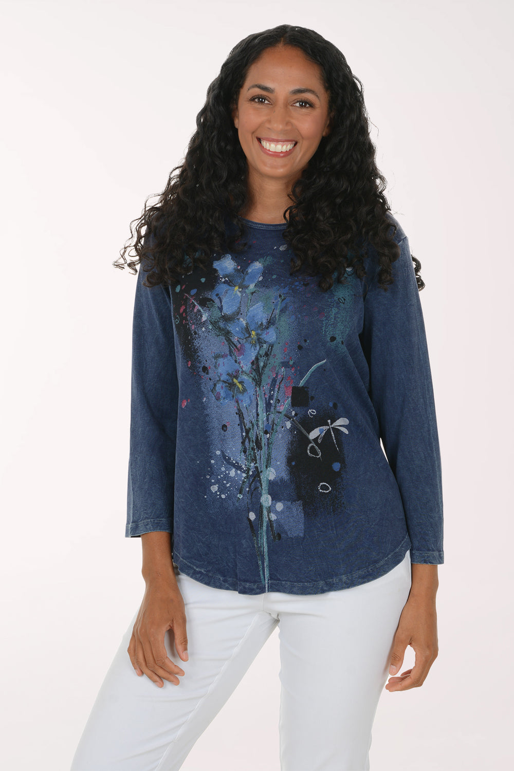 A woman with long curly black hair wearing a blue long-sleeve top with a colorful abstract floral print and white pants, standing against a plain white studio background.