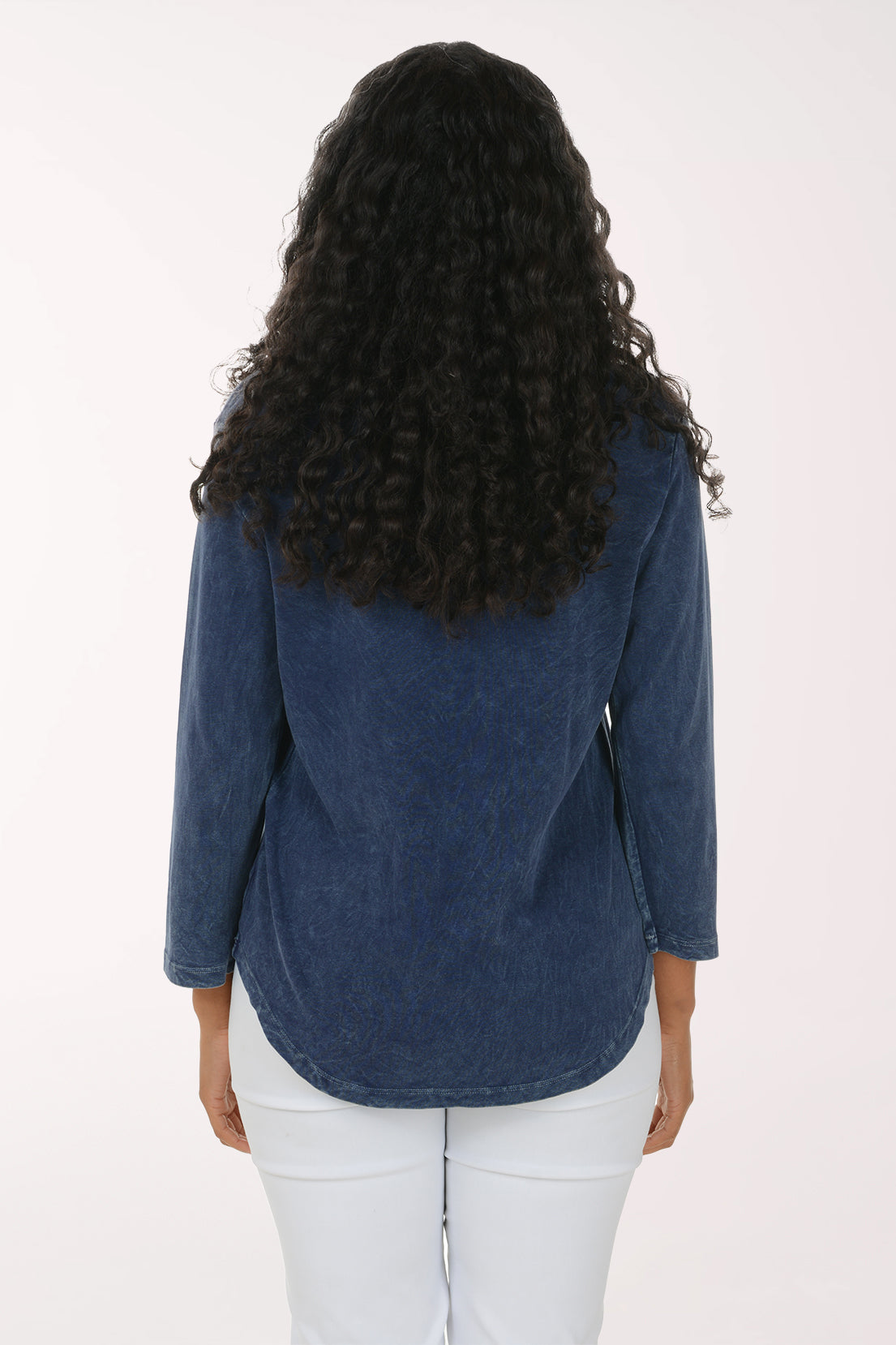A woman with long curly black hair wearing a blue long-sleeve top with a colorful abstract floral print and white pants, standing against a plain white studio background.