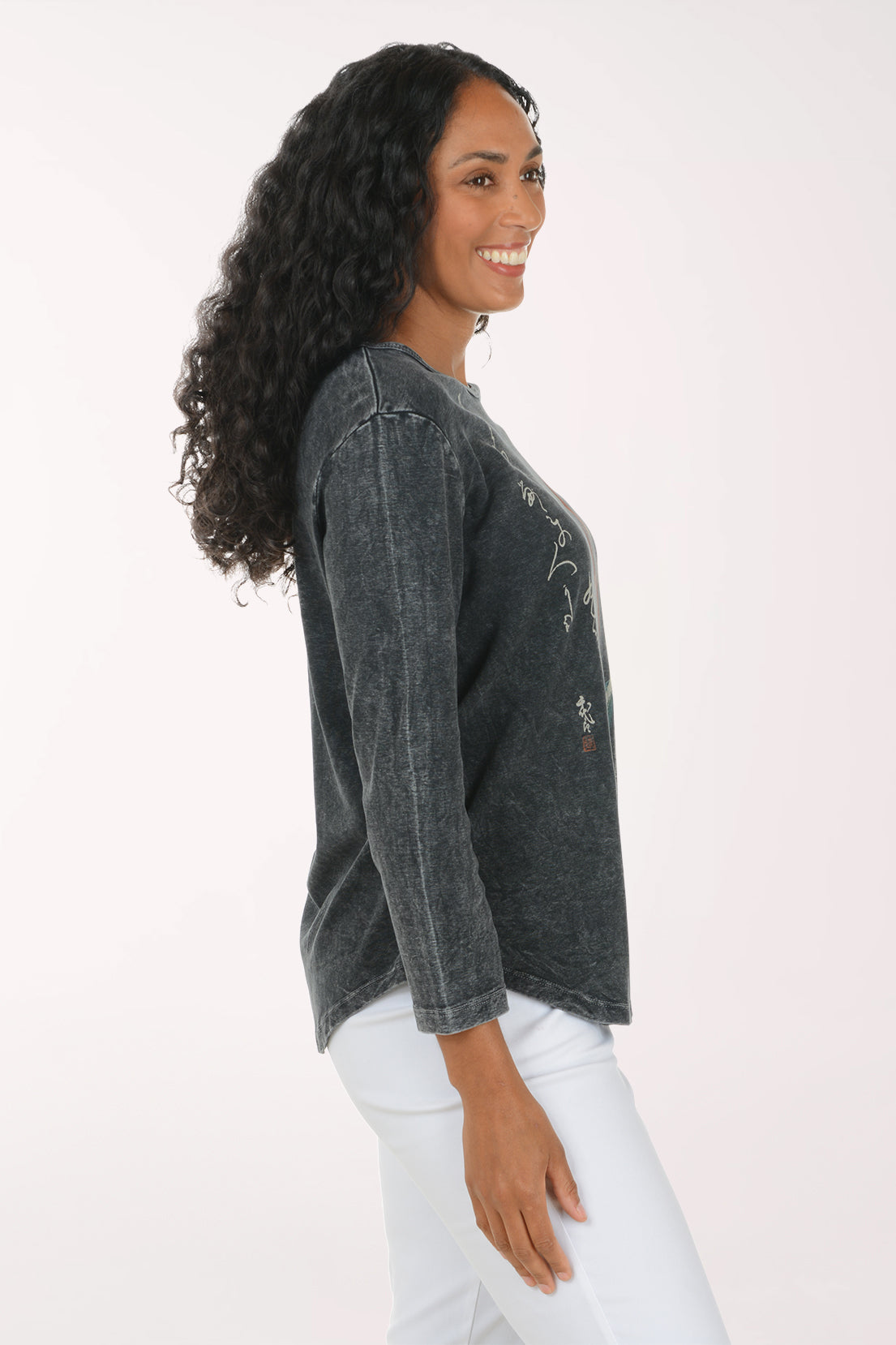 A woman with long, curly black hair is shown from a side profile, wearing a dark gray mineral-wash long-sleeve top with white embroidery and white pants, set against a plain white studio background.