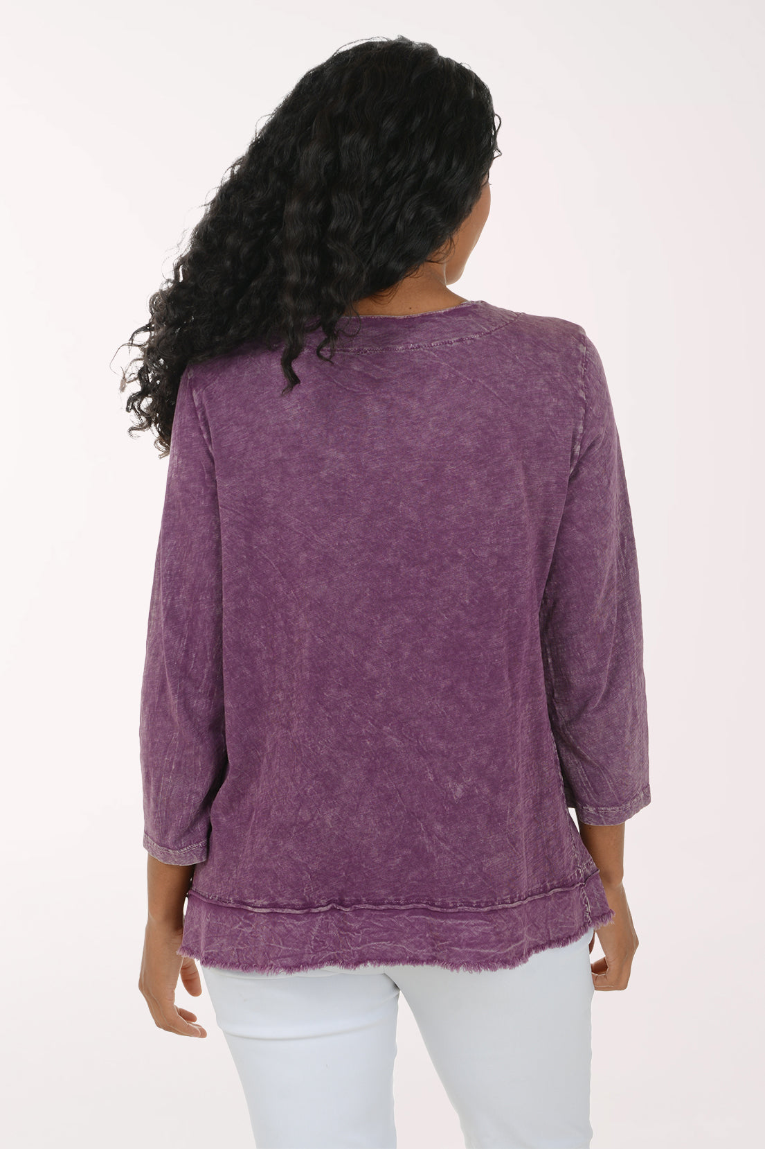 A woman modeling a purple, long-sleeved, acid-wash top with a colorful abstract floral graphic on the front, paired with white pants, against a plain white background.