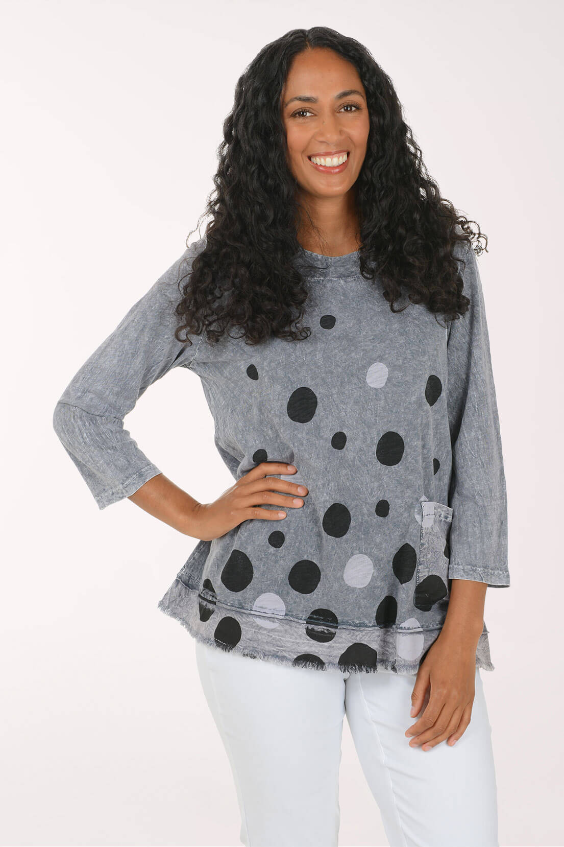 A woman with long curly black hair wearing a gray three-quarter sleeve top with a black and white polka dot pattern, paired with white pants, posing against a plain white background.