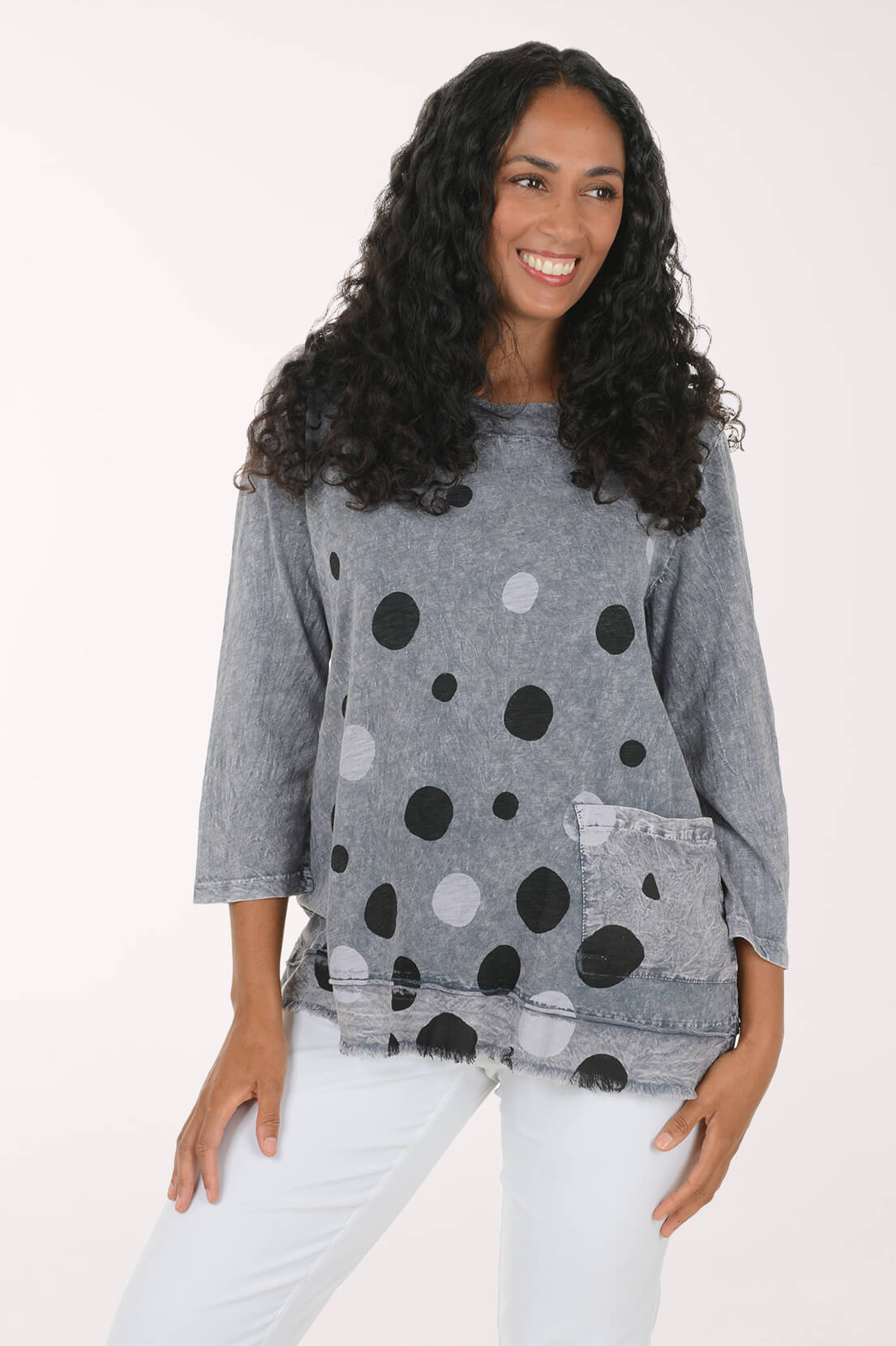 A woman with long curly black hair wearing a gray three-quarter sleeve top with black and white polka dots and white pants, posing against a solid white studio background.