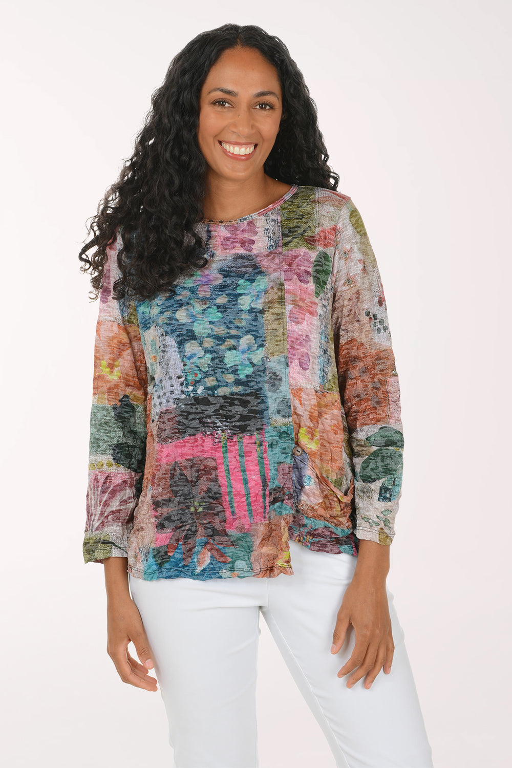 A woman with long curly black hair wearing a colorful abstract patterned long-sleeve top and white pants, standing against a plain white studio background.