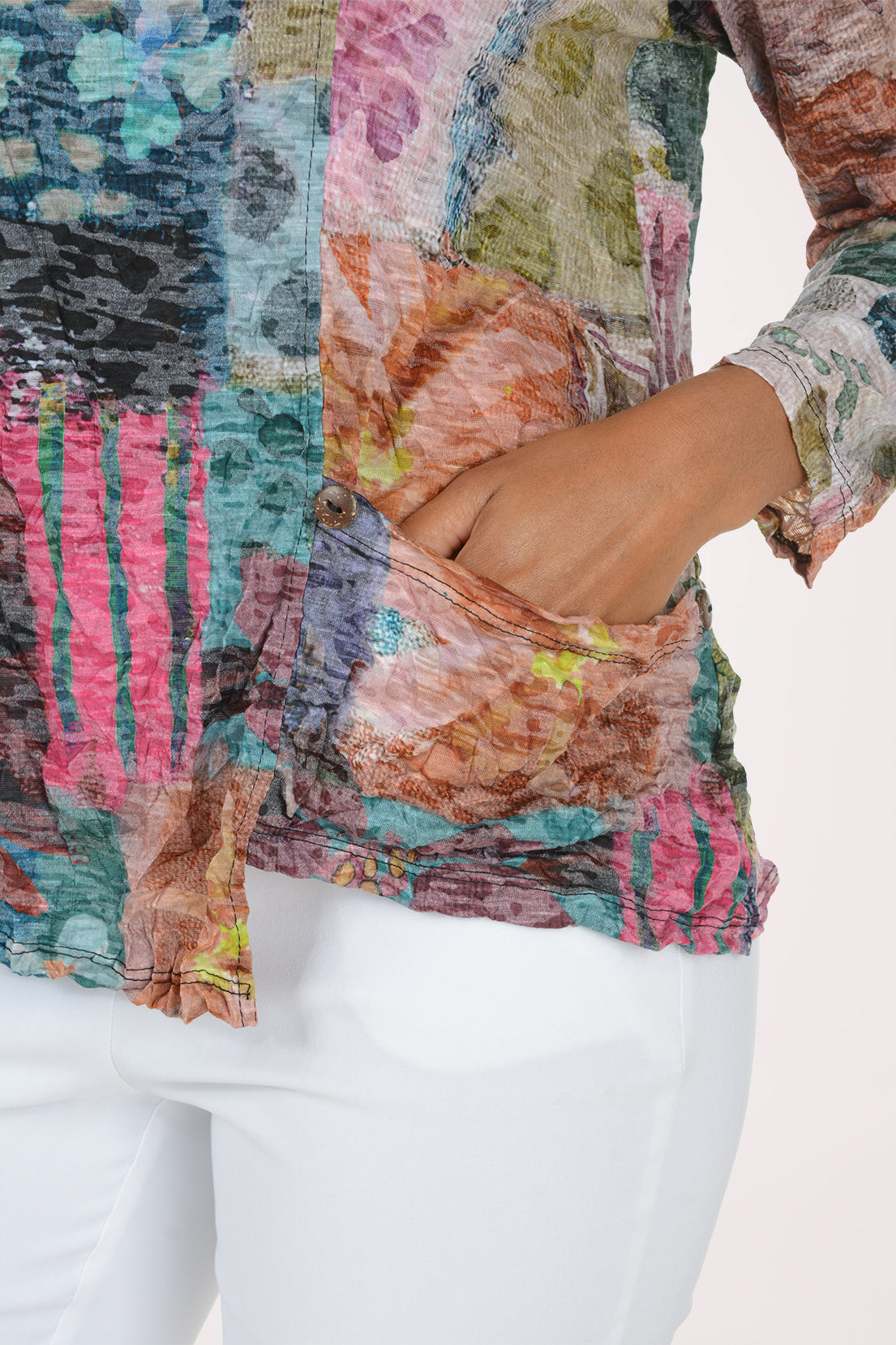 A close-up of a woman wearing a multicolor patchwork print top with a side pocket, shown from the chest down with white pants against a plain light background.