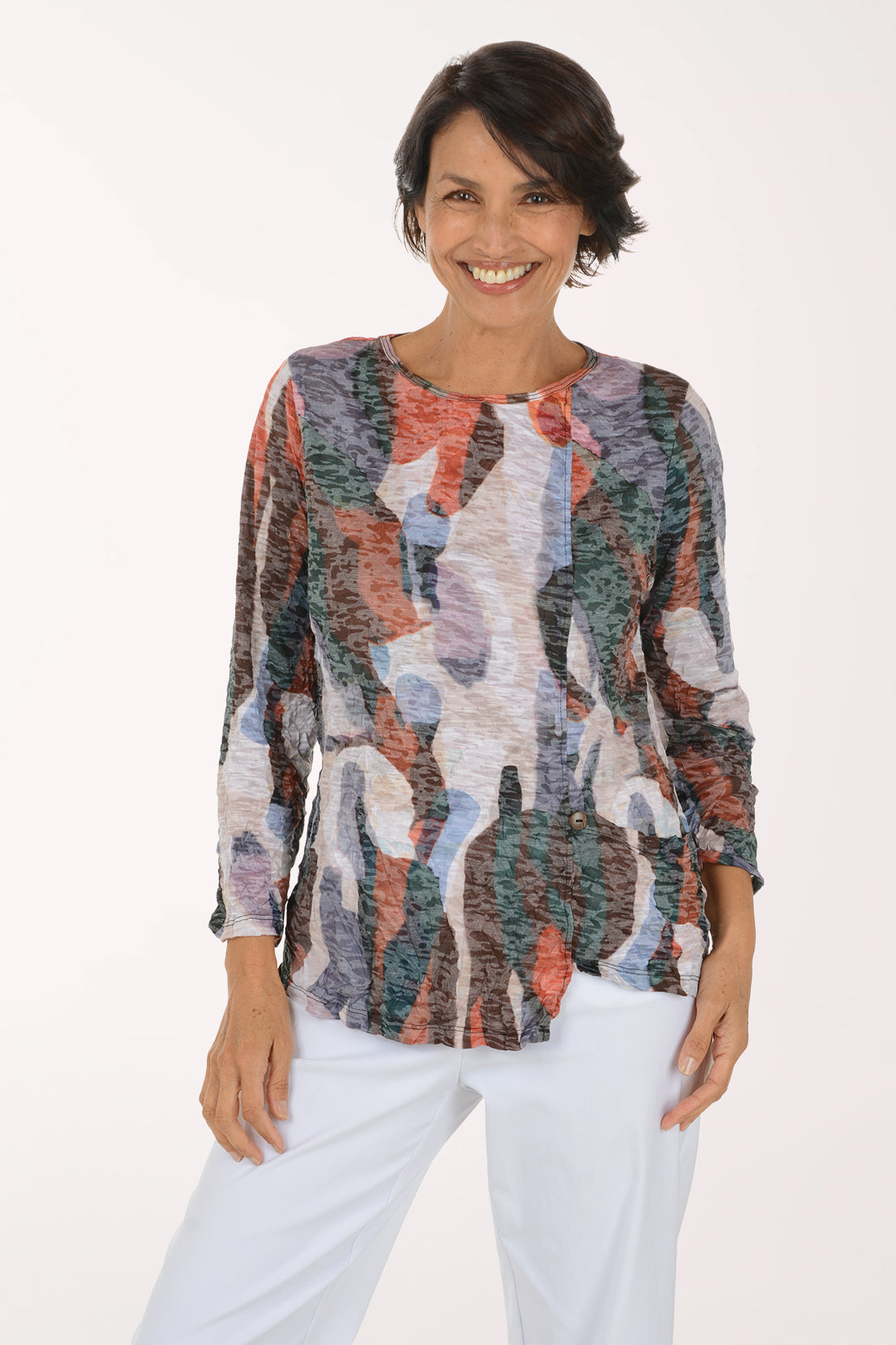 A smiling woman wearing a long-sleeve abstract print top with a mix of orange, blue, green, gray, and white tones, paired with white pants, against a plain white studio background.