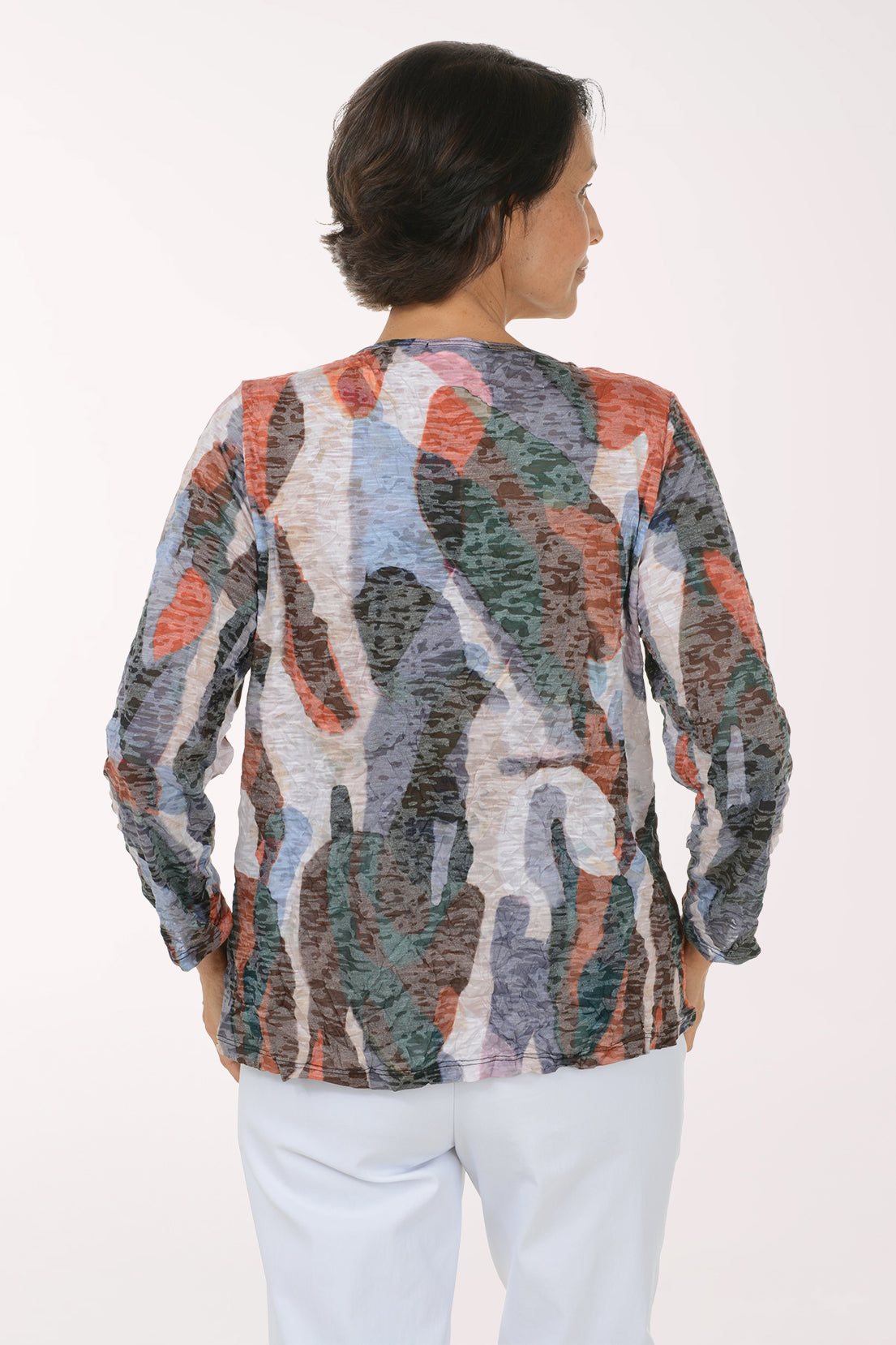 A woman viewed from the back wearing a long-sleeve abstract patterned top with a mix of gray, orange, blue, green, and white tones, paired with light blue pants, against a plain white background.