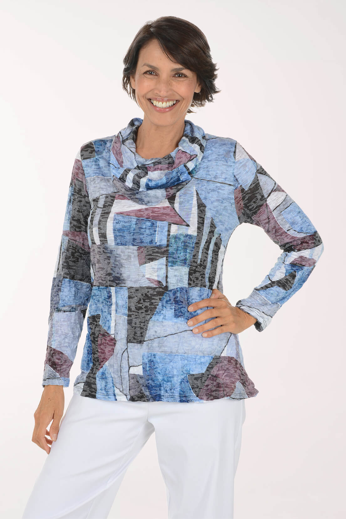 A smiling woman wearing a long-sleeve abstract print top with a cowl neck in shades of blue, gray, and mauve, paired with white pants, against a plain white background.