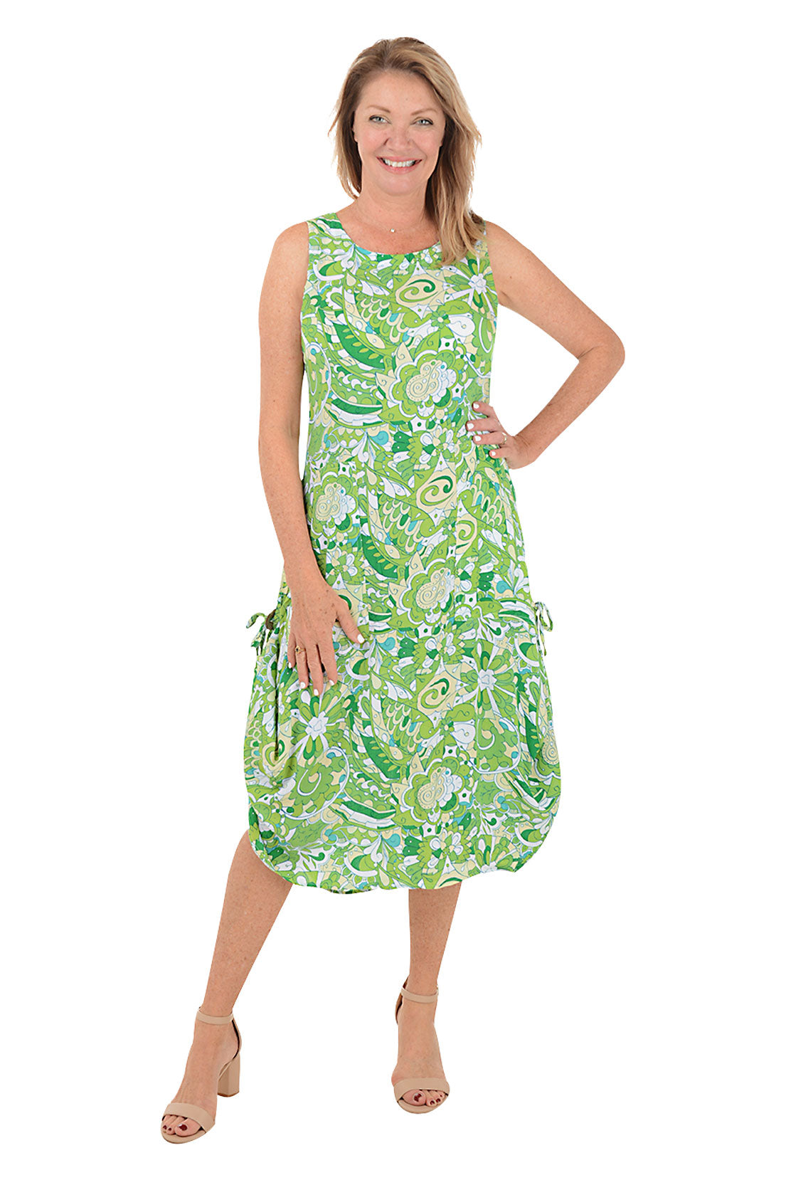 A front shot of a model wearing a sleeveless, knee-length, green and white paisley print dress with a scoop neckline, dual front patch pockets, and a shark bite hemline.
