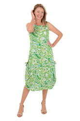 A front shot of a model wearing a sleeveless, knee-length, green and white paisley print dress with a scoop neckline, dual front patch pockets, and a shark bite hemline.
