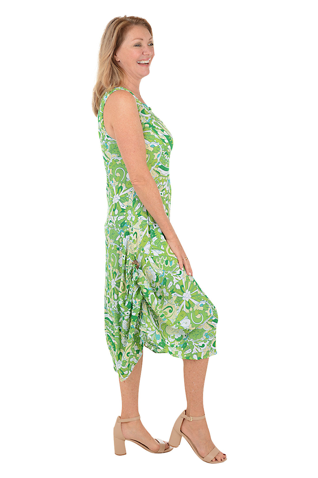 A side shot of a model wearing a sleeveless, knee-length, green and white paisley print dress with a scoop neckline, dual front patch pockets, and a shark bite hemline.