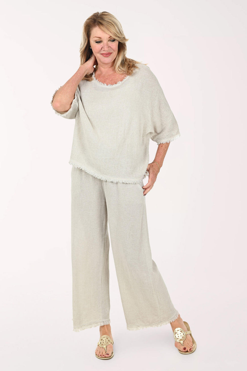 A woman modeling a light beige two-piece lounge set consisting of a short-sleeve top with a raw hem and matching wide-leg pants, styled with gold sandals against a plain white background.