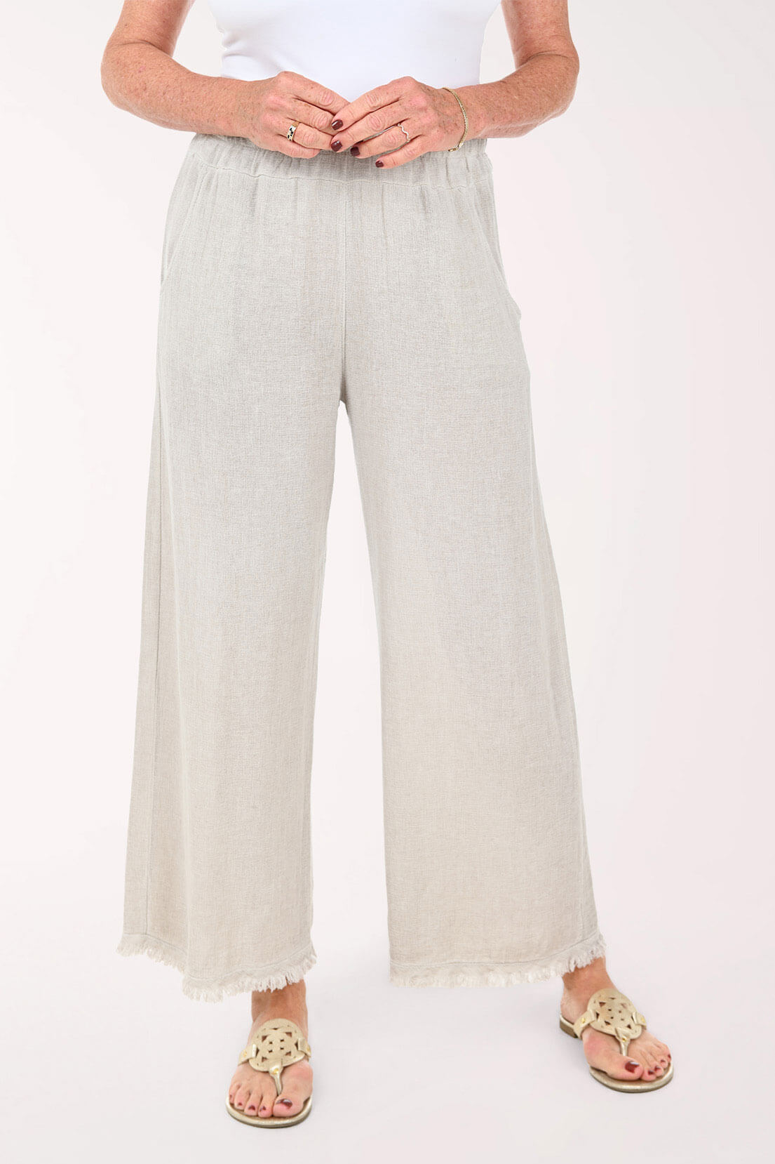 A person wearing light beige wide-leg cropped pants with a subtle textured fabric and frayed hems, paired with a white top and tan sandals, shown against a plain light background.