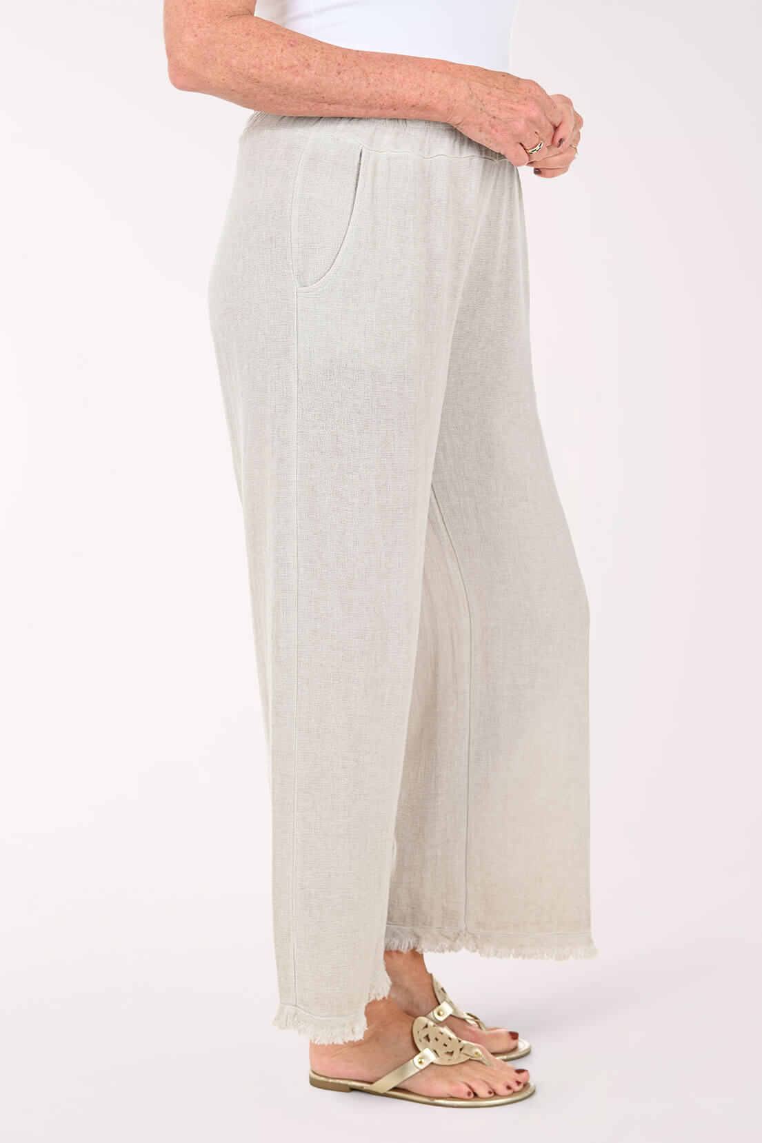 A person wearing light beige wide-leg cropped pants with a subtle textured fabric and frayed hems, paired with a white top and tan sandals, shown against a plain light background - side shot