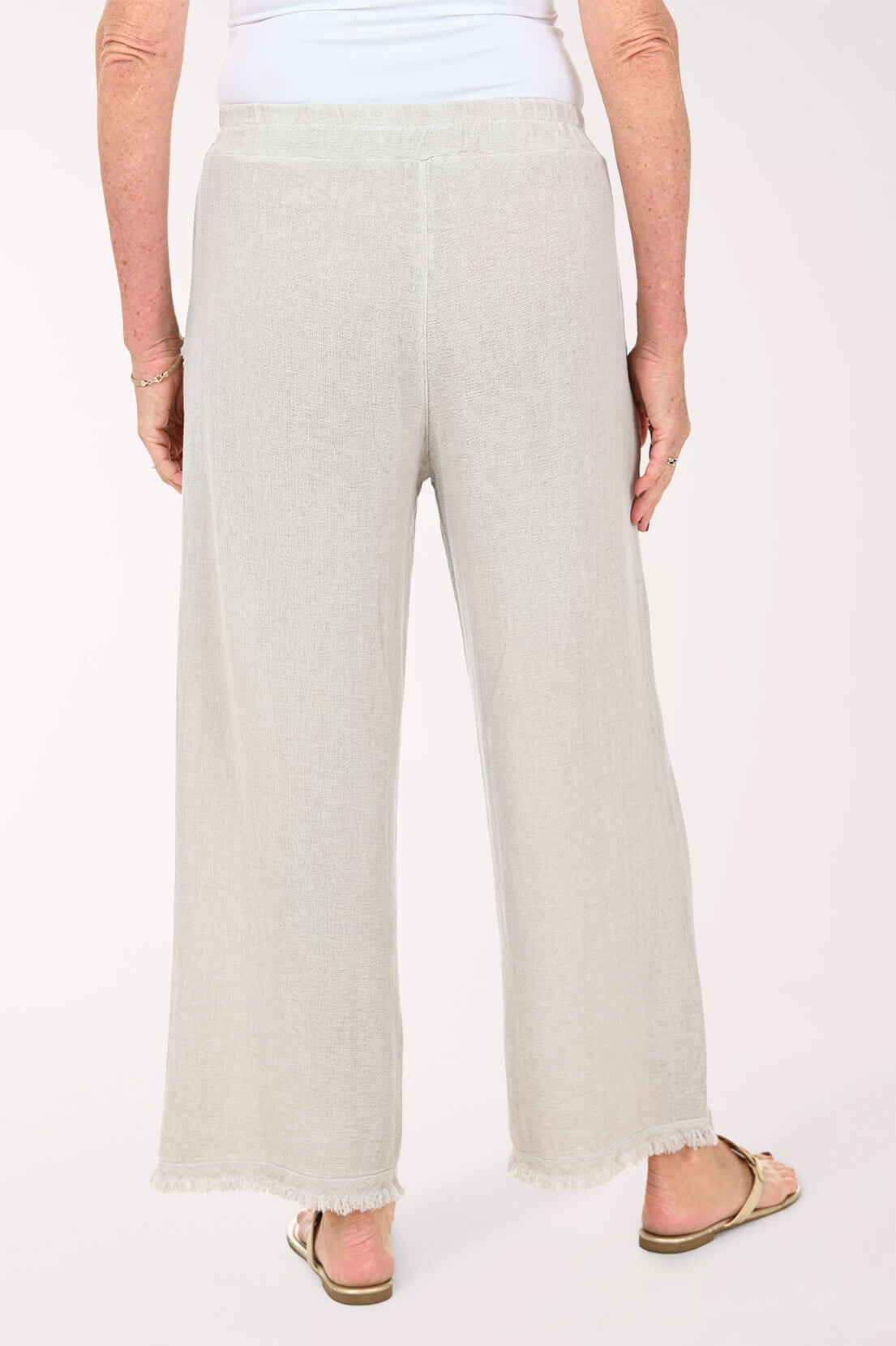 A person shown from the back wearing light beige wide-leg cropped pants with a frayed hem, paired with a white top and tan sandals, against a plain light background.
