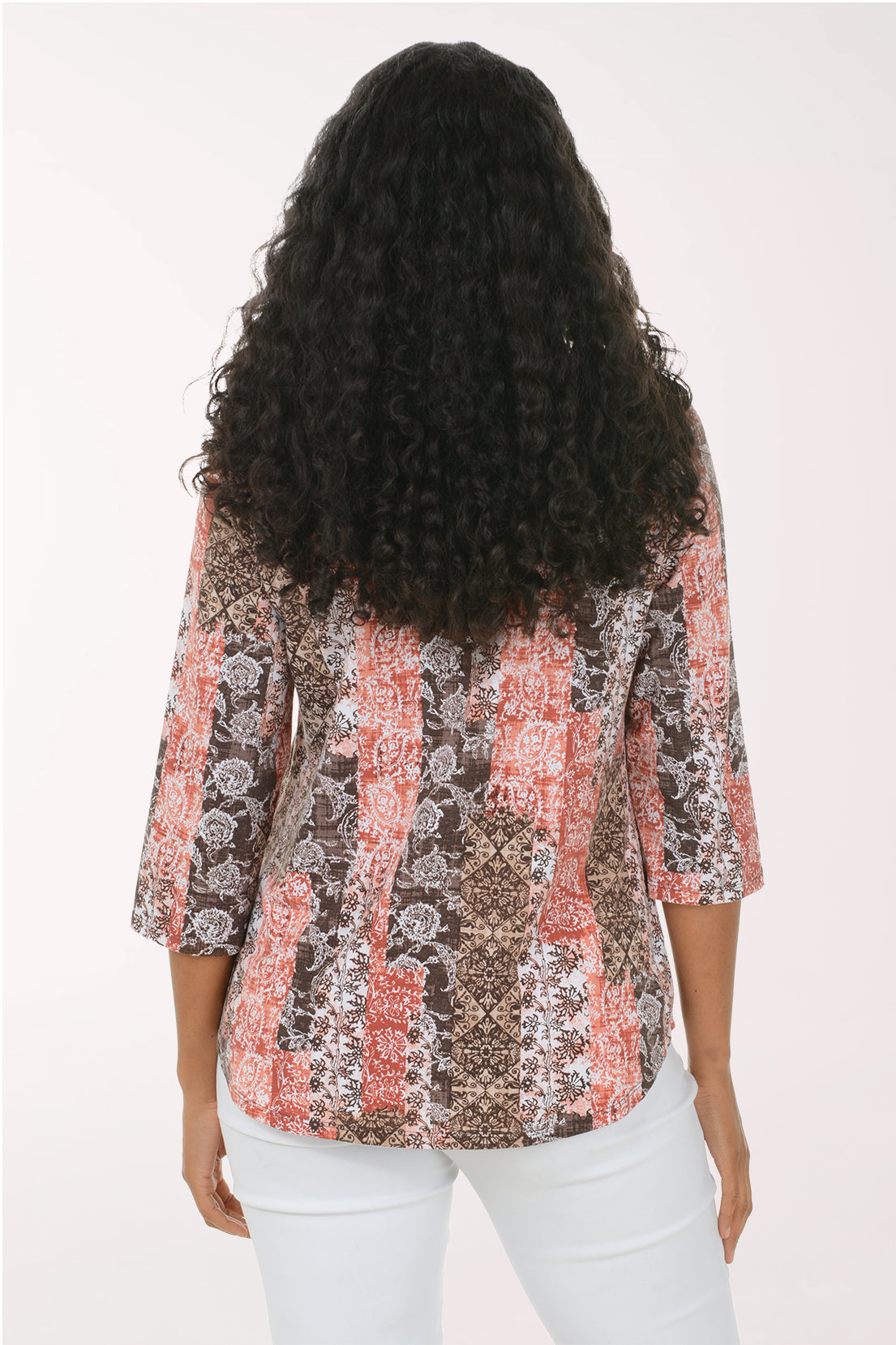 A woman with long curly black hair wearing a three-quarter sleeve patterned blouse with white pants, standing against a plain white studio background.